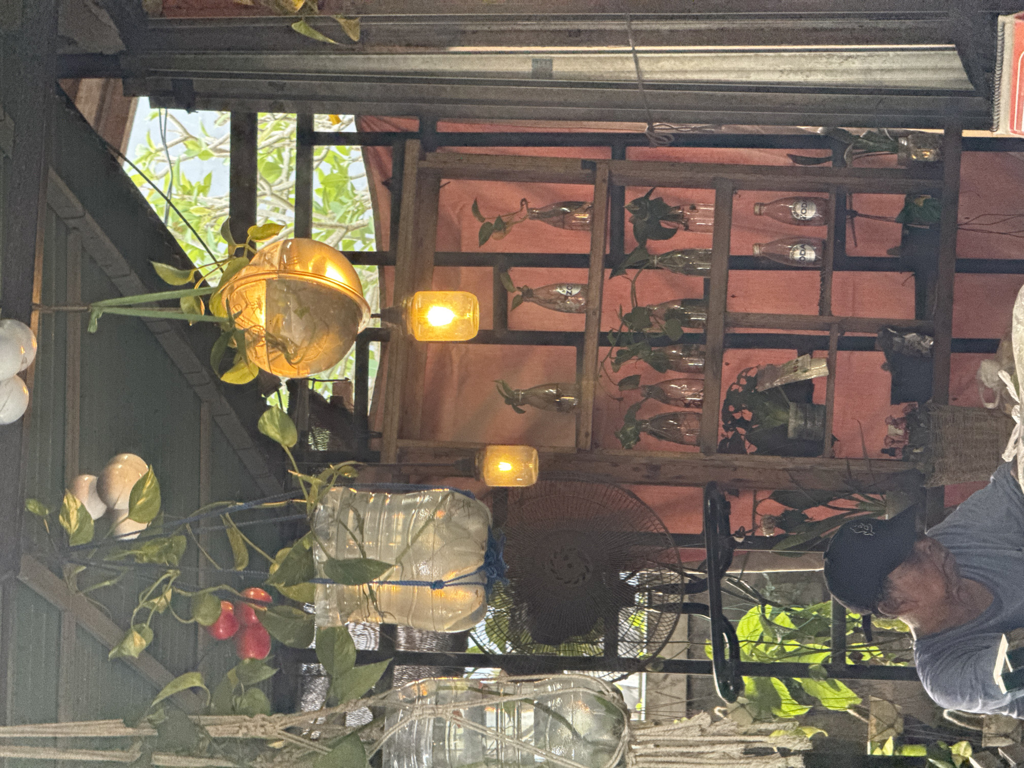 The image captures a dimly lit, bohemian-style cafe interior, likely in Kuta Selatan, Indonesia. Sunlight filters through the open-air structure, illuminating hanging plants in repurposed plastic bottles and a globe-shaped planter. A rustic wooden shelving unit displays various glass bottles, some containing plant cuttings, and two vintage "Soda" bottles. A vintage oscillating fan is visible in the background. In the foreground, an elderly man wearing a dark baseball cap and a grey t-shirt sits, appearing to be engrossed in something on the table in front of him, out of frame. The overall ambiance is relaxed and eclectic, with a clear emphasis on natural elements and repurposed materials.