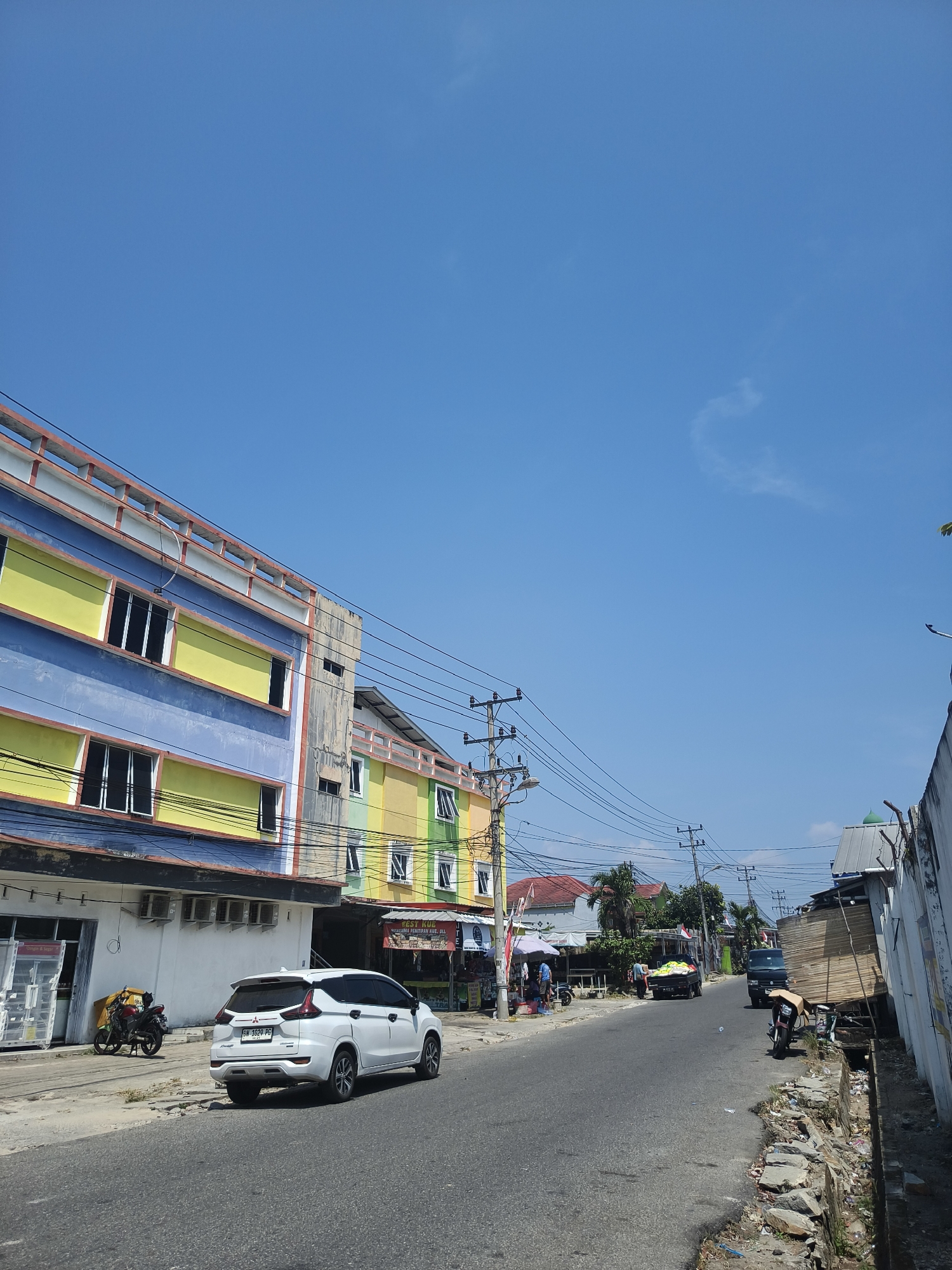 The image depicts a street scene in Pangkalpinang, Indonesia, under a clear blue sky. On the left side of the road, a multi-story building with brightly colored facade in blue, yellow, and red stands prominently. Several windows are visible, some open and some closed. Air conditioning units are mounted on the building's facade. A white car, a Mitsubishi Xpander with license plate BN 1020 PG, is parked on the side of the road, facing away from the camera. Next to it, a red motorcycle is parked. Power lines crisscross the sky above the street, supported by utility poles.
Further down the street, more buildings with similarly vibrant colors, including green and yellow, line the right side. Various stalls and shops are present, some with awnings. A black truck loaded with what appears to be lumber is parked on the right side of the road, with a person standing near it. Another black truck is further down the road. People can be seen walking and interacting near the shops. The street itself is paved, with a damaged concrete drainage channel on the right side, containing debris. The overall impression is a bustling commercial street on a sunny day.