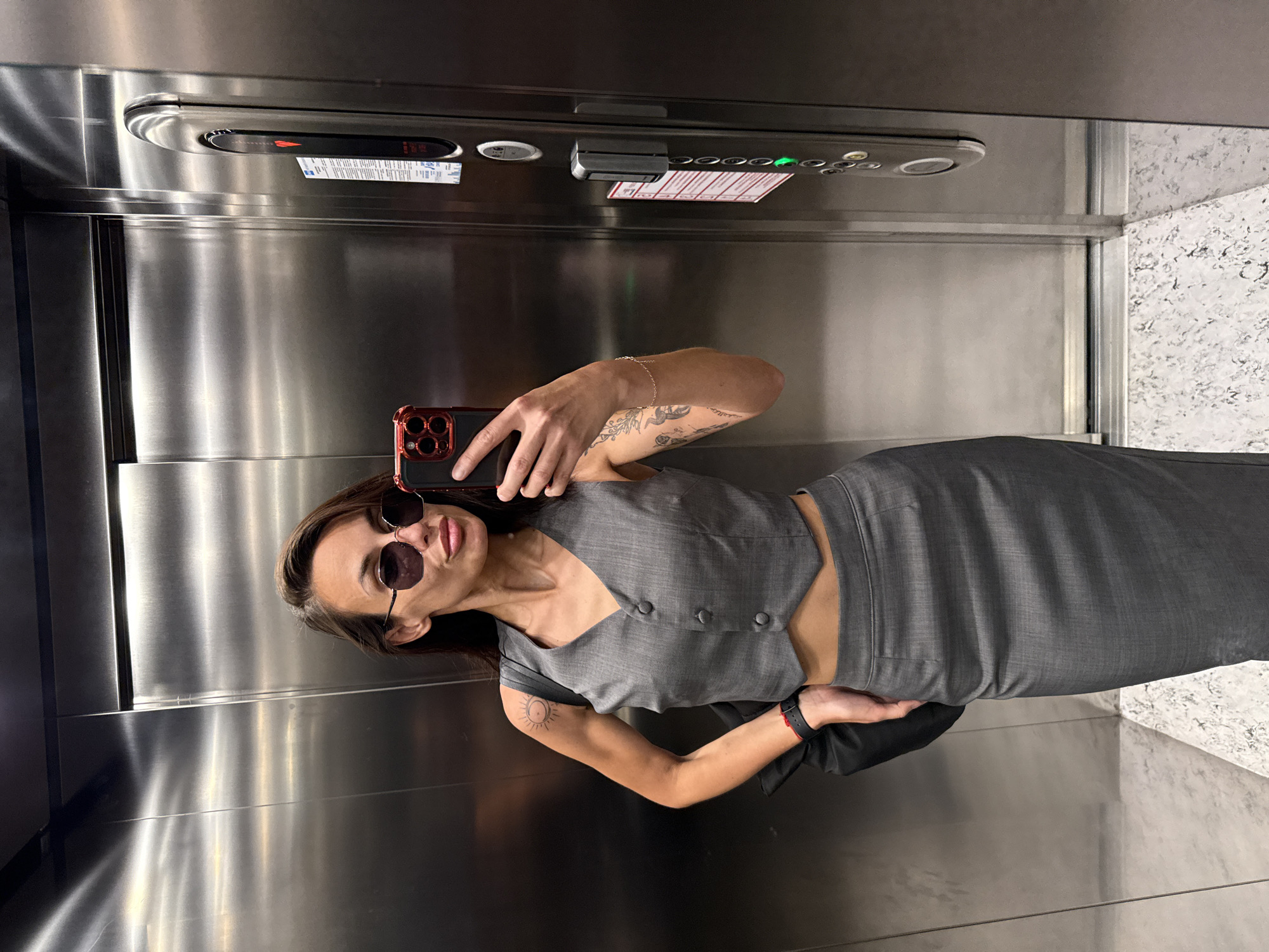 A woman stands inside an elevator, taking a selfie. She wears sunglasses, a gray vest, and a matching skirt, showing a small portion of her midriff. She has tattoos on her arms. She holds a smartphone with a red case. The elevator interior is stainless steel, with a control panel visible at the top of the frame. Part of the elevator floor and wall, which appear to be a marble-like material, are visible on the right. The location is likely a building in a city, potentially Pleven, Bulgaria, but the specific location within the city is not discernable from the image.