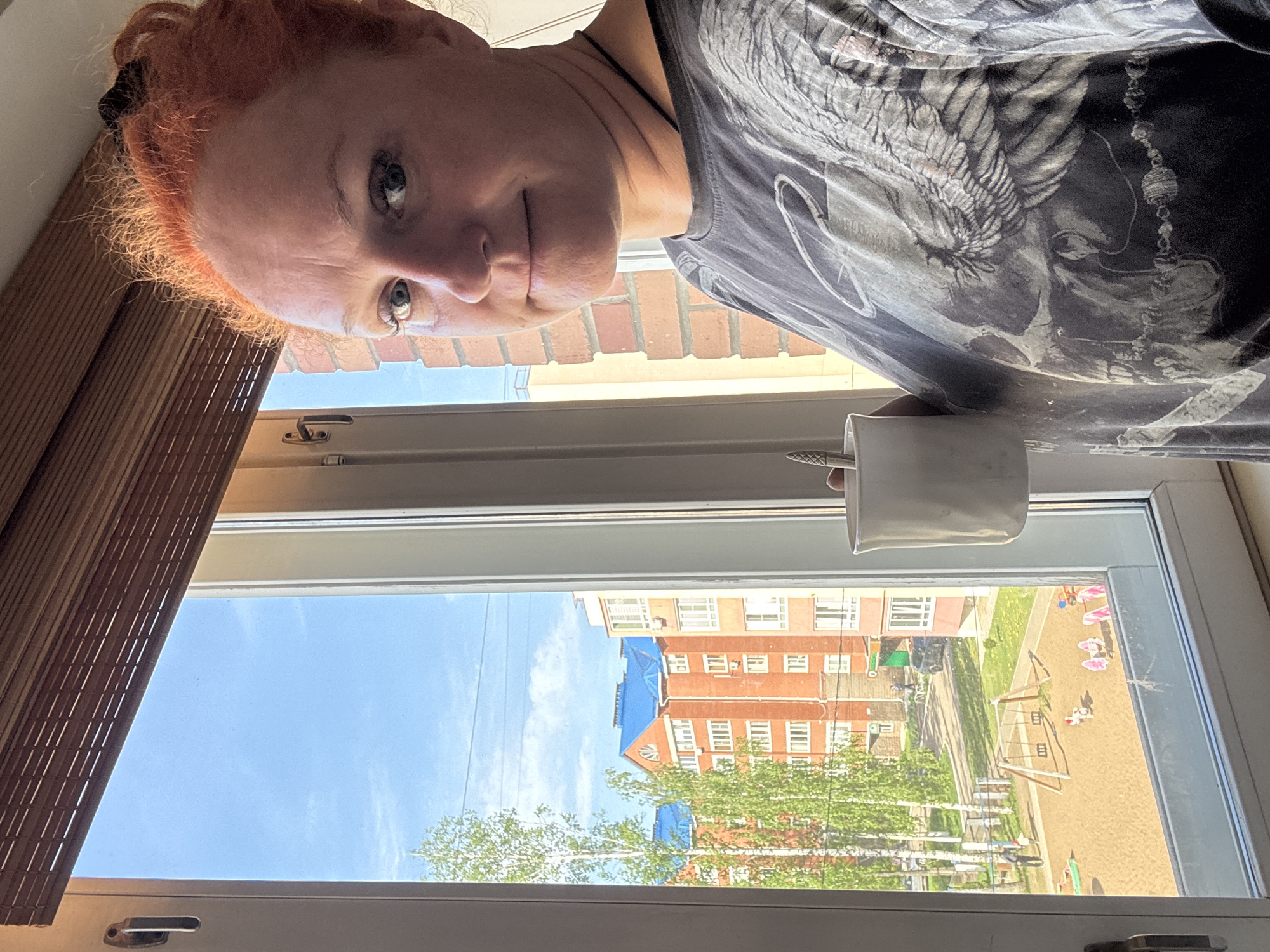 A selfie of a woman with red hair looking down at the camera. She is wearing a grey t-shirt with a dark print. She is holding a white mug with a spoon in it. Behind her is an open window looking out onto a view of a residential area in Chaikovsky, Russia. The view includes a brick building with blue tarp covering part of the roof and a playground with several swings. The sky is blue and clear. A partial view of wooden blinds is seen on the left of the frame.