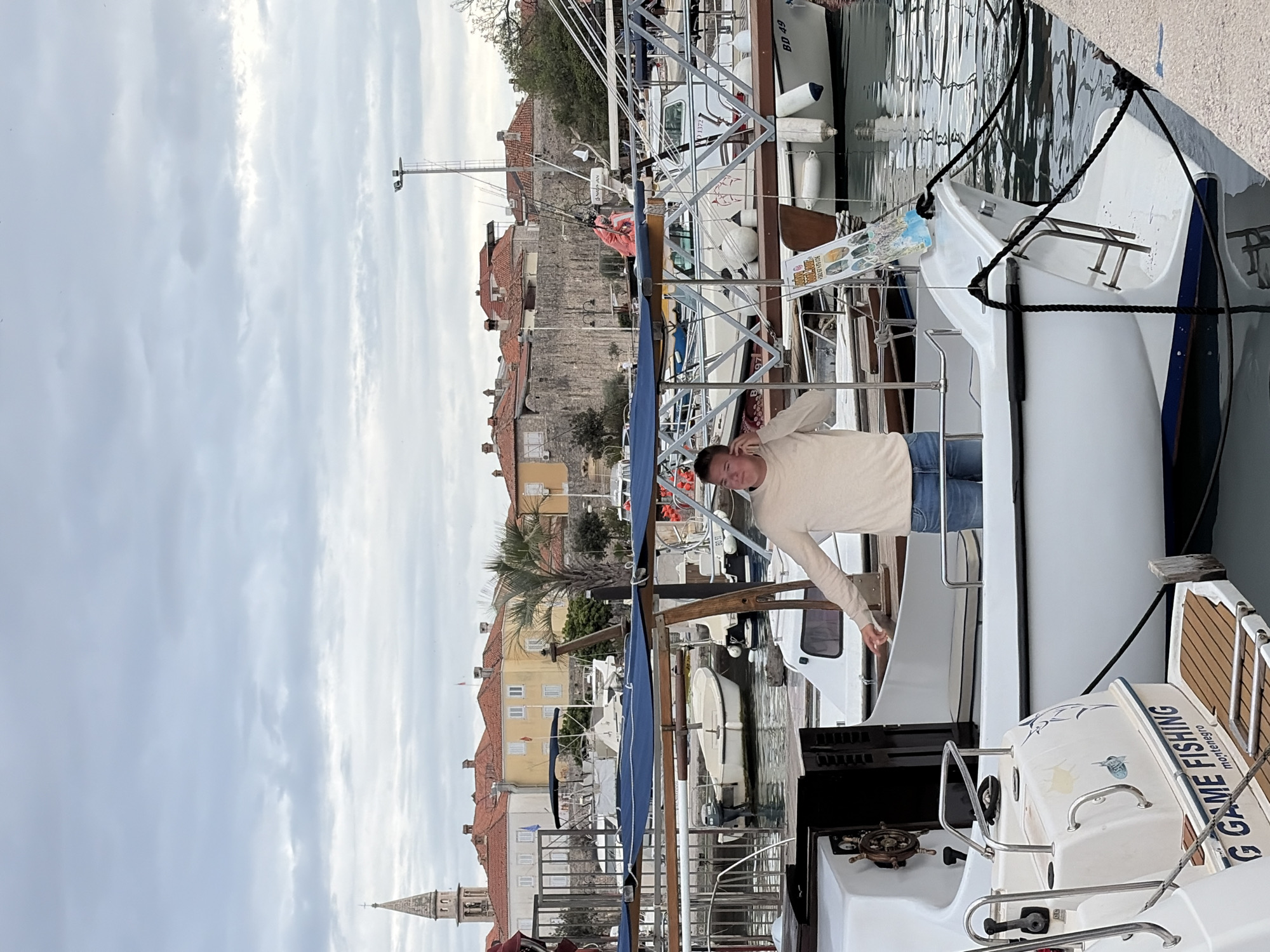 The image shows a man standing on a boat in a marina. The buildings of Budva, Montenegro, are visible in the background. The sky is overcast. The man is wearing a beige sweater and blue jeans. He has his arms outstretched. The boat has the words "GAME FISHING" printed on it. Other boats are moored nearby.