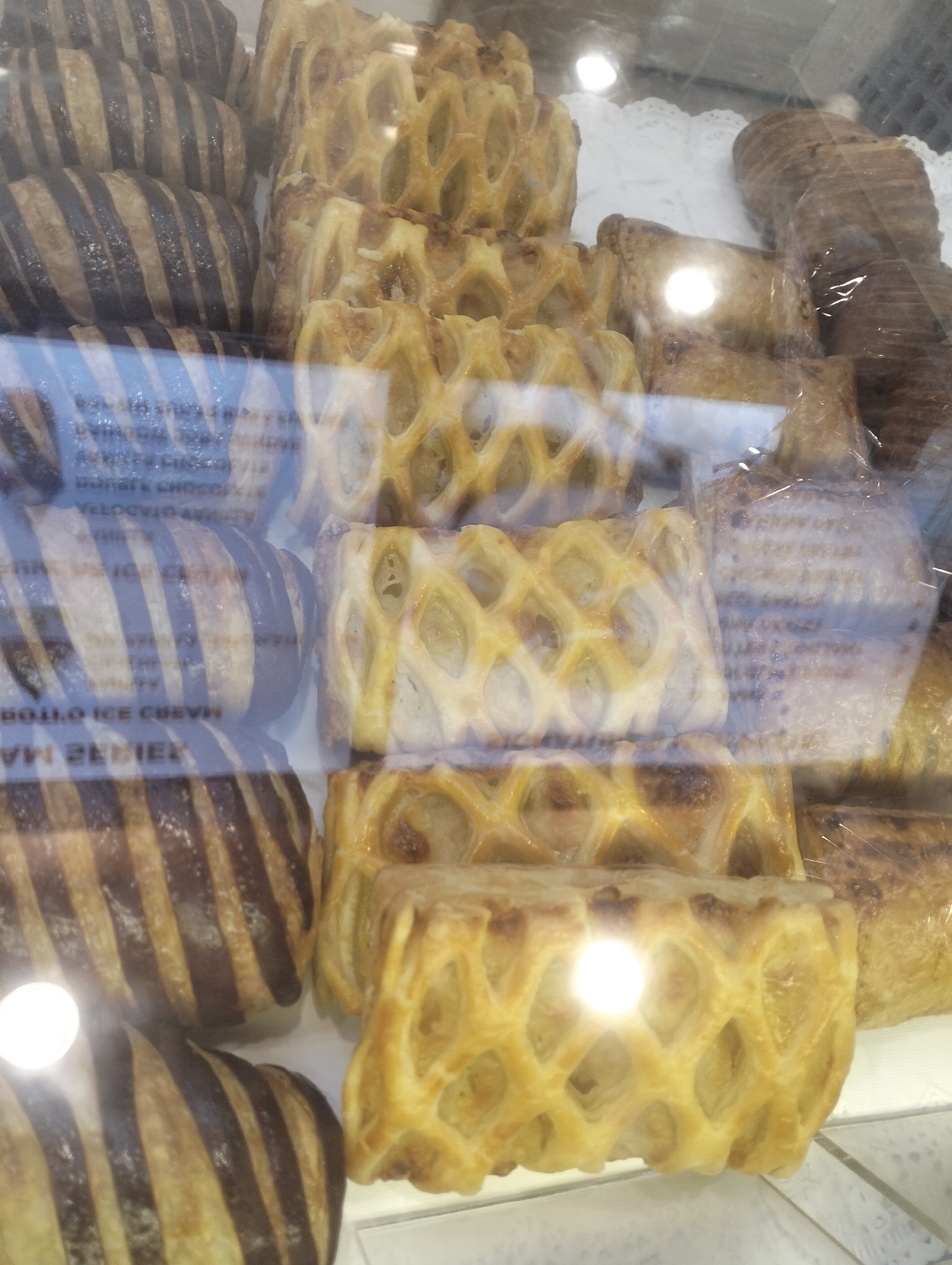 The image displays a close-up view of a bakery display case filled with various pastries. In the foreground, several chocolate-striped pastries are arranged. To the right and behind these, there are lattice-topped pastries with visible filling, and further back, more pastries wrapped in plastic. The lighting in the display case creates bright reflections on the glass and the pastries themselves, obscuring some details. Text is visible through the glass, suggesting labels or signage for the products. The overall impression is one of a well-stocked bakery counter. There are no people visible in the image, and no specific details about the location in Air Jamban, Indonesia, can be inferred from the visual content alone.