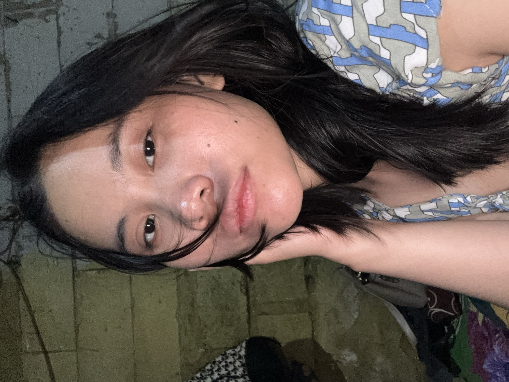 The image is a close-up, medium shot of a young woman with long, dark hair. She is looking towards the camera with her right hand supporting her jaw. Her hair is partially obscuring her face, with some strands falling across her eyes and cheek. She is wearing a patterned shirt with blue, white, and grey designs. The background appears to be a rough, textured wall, possibly made of concrete blocks, with some items of clothing and bags visible to the left of the woman. The lighting is dim, casting shadows and highlighting parts of her face. The scene appears to be indoors, in what looks like a dimly lit room or an outdoor shelter, consistent with a residential area in East Jakarta, Indonesia.
