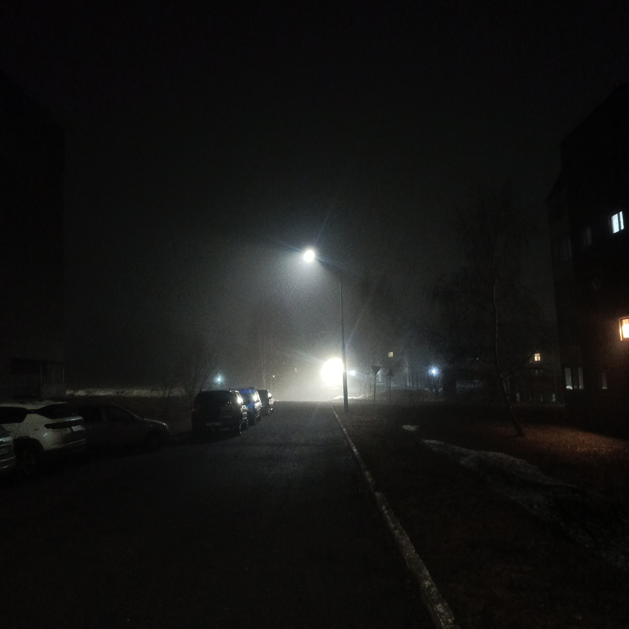 The image depicts a dimly lit street scene in Марковский, Russia, at night. Heavy fog obscures visibility. A row of parked cars lines the left side of the street, illuminated by the headlights of a vehicle further down the road. A street light emits a strong glow, contributing to the hazy atmosphere. On the right side of the street, a grassy area with patches of melting snow is visible. Buildings flank both sides of the street, with faint lights visible in some windows on the right. The street surface appears wet and reflective. There are no people visible in the image.