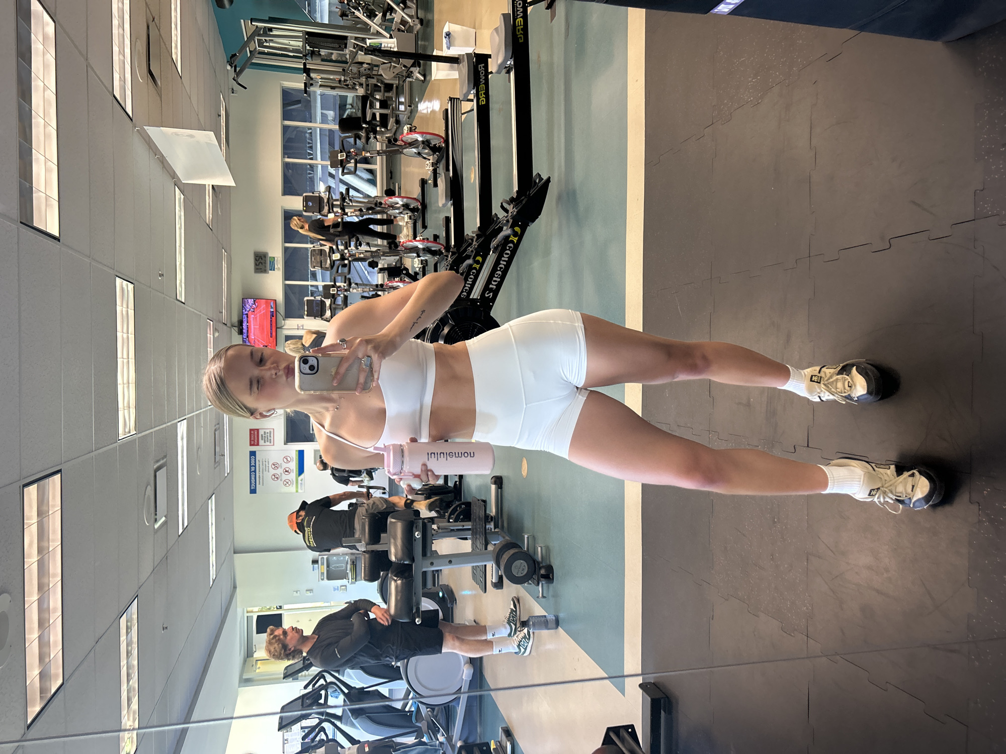 The image is a selfie taken in a gym that appears to be in Whistler, Canada. The person taking the photo is a young woman in a white sports bra and shorts. She is holding a pink water bottle and using her phone to take the picture in front of a mirror. In the background, there are various pieces of gym equipment, including rowing machines, weight machines, and treadmills. There are other people in the gym, some of whom are using the equipment. The gym has a light blue and white color scheme with black rubber flooring. The lighting is bright, suggesting a well-lit interior space.