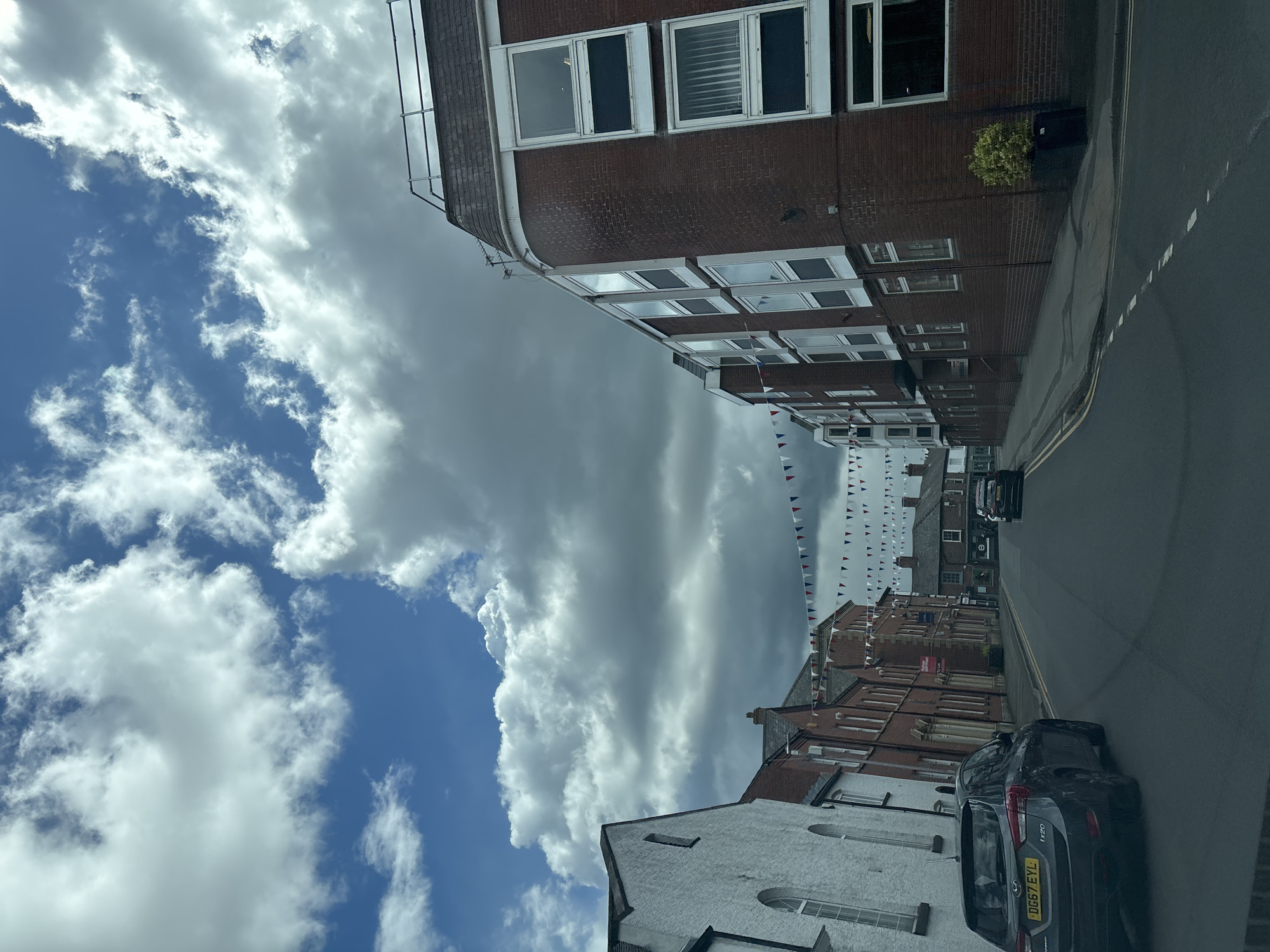 The image shows a street scene in Knutsford, United Kingdom. The view is of a street lined with buildings of varying heights and architectural styles, a mix of brick and white-painted facades. Bunting is strung across the street. The sky is filled with both blue sky with scattered white clouds and a large mass of grey storm clouds. A dark colored vehicle is parked on the right side of the street in the foreground. Another dark colored vehicle is further down the street. The street is paved and has painted yellow lines denoting parking areas.