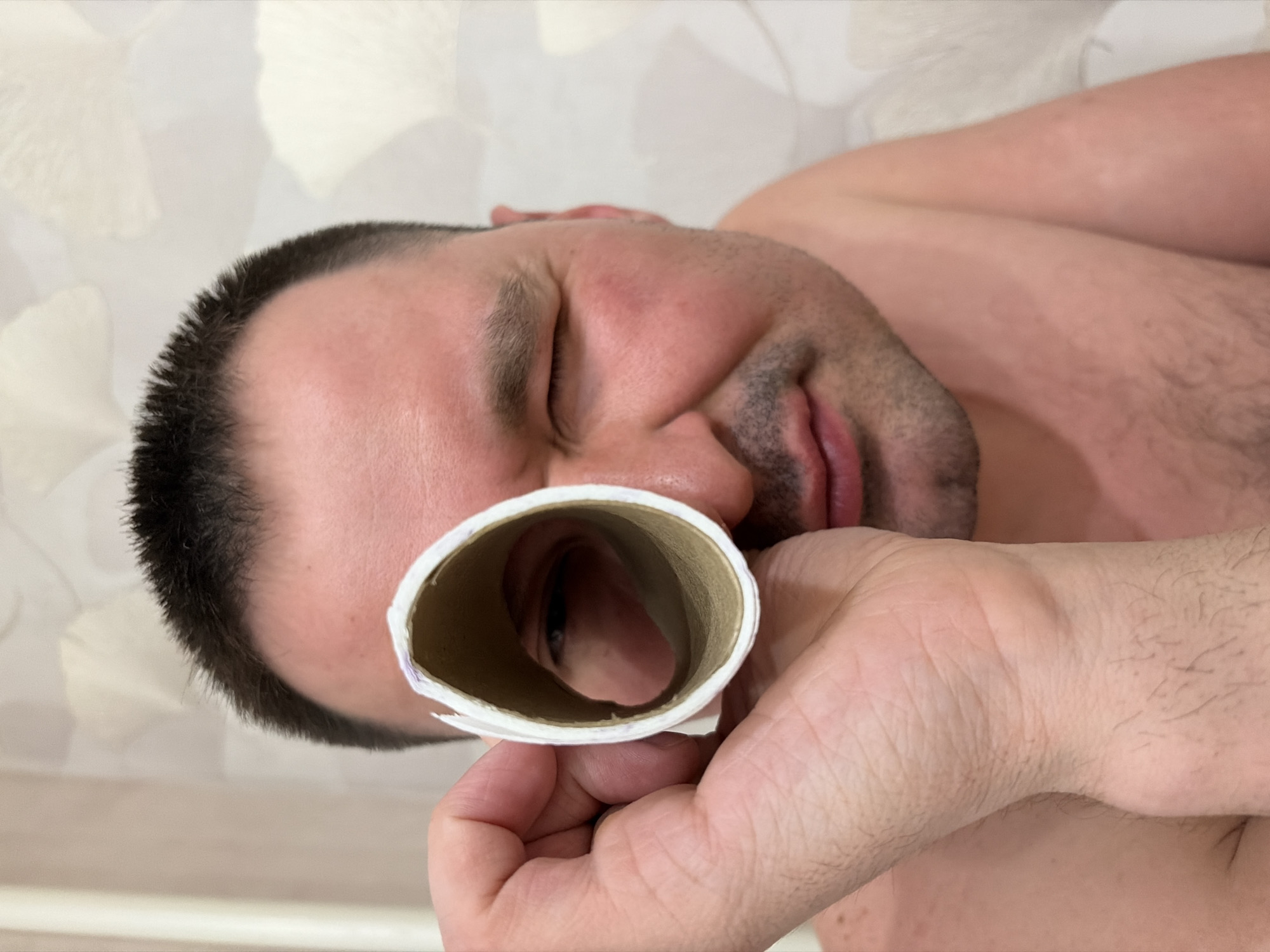 A man is holding an empty toilet paper roll in front of his eye, looking through it. He is lying down, shirtless, with his eyes partially closed. The background shows a light-colored wall with a faint pattern. The man has short dark hair and a stubbly beard. There is no indication that this photo was taken in Izhevsk, Russia.