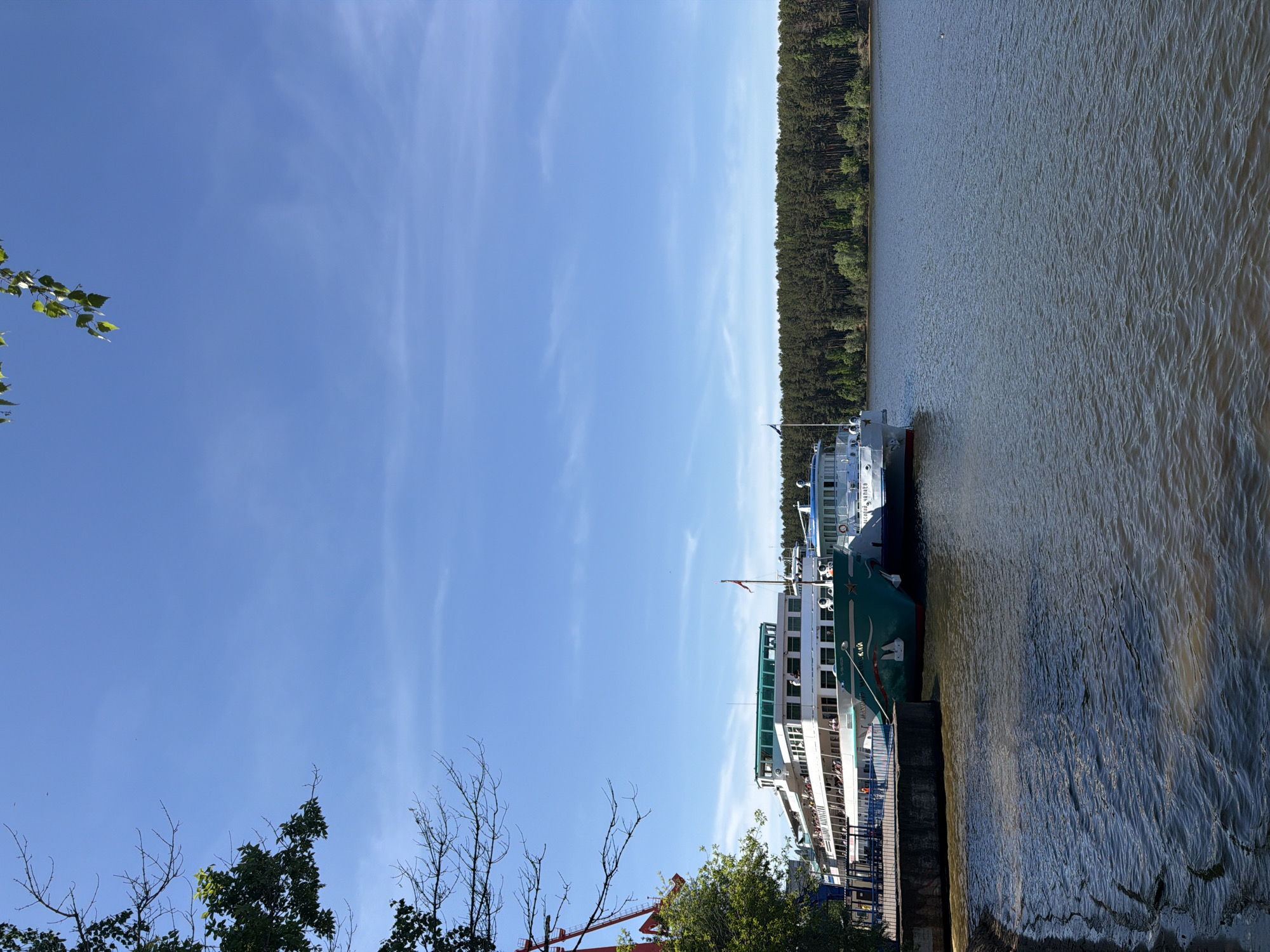A daytime shot of a river with two passenger ships docked on the right side of the frame. A strip of forest lines the opposite bank. To the left, a blue sky with thin, wispy clouds dominates the frame. In the foreground, trees and a red crane are visible.
The location is Chaikovsky, Russia. The passenger ships are docked, presumably for passengers to board or disembark. A crane suggests construction or maintenance activities near the dock. The weather appears clear and sunny.