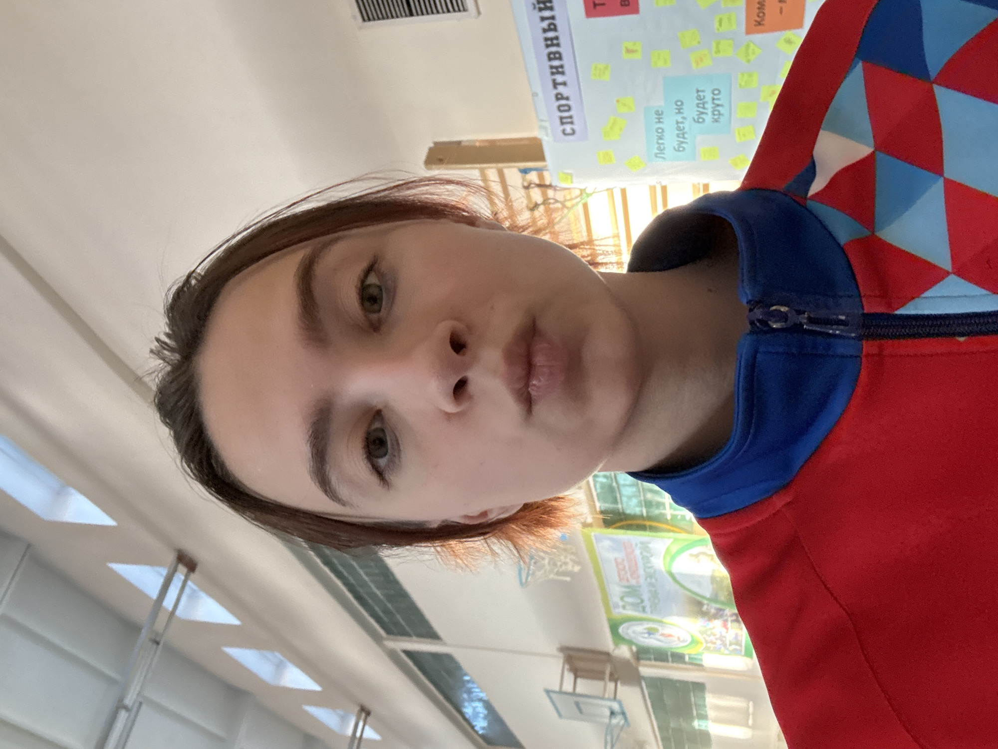 A young person is taking a selfie inside what appears to be a school gymnasium in Berezniki, Russia. The person has light skin, brown hair, and is wearing a red jacket with blue accents around the neck and shoulders. In the background, there is a wall with a poster that says "Спортивный" (Sportive) in Russian and some post-it notes. Further in the background, there is a basketball hoop and windows with metal frames. The gym seems well-lit.