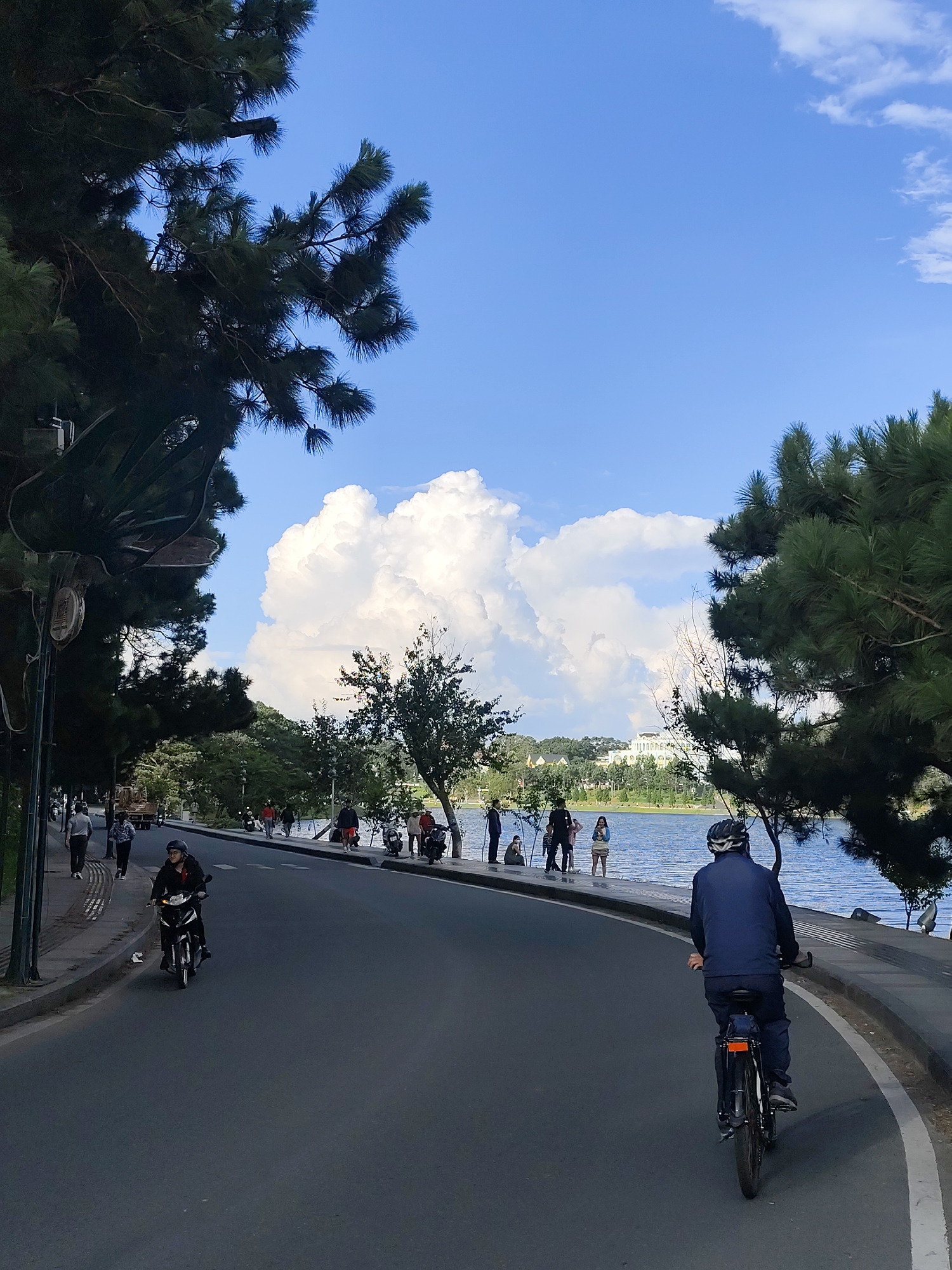 This image depicts a sunny day by Xuan Huong Lake in Da Lat, Vietnam. A paved road curves along the lakeside, with a pedestrian walkway on the left. Several individuals are present: a person rides a motorcycle in the foreground on the left, while a cyclist in a blue jacket and helmet is further ahead on the right. On the walkway, a group of people are gathered, some standing and others sitting. Pine trees line the road and the lake, casting shadows. The lake is calm and reflects the bright blue sky, which is dotted with large, fluffy white clouds. In the distance, a white building with classical architecture is visible on the far shore, surrounded by trees. The overall scene conveys a peaceful and active atmosphere.