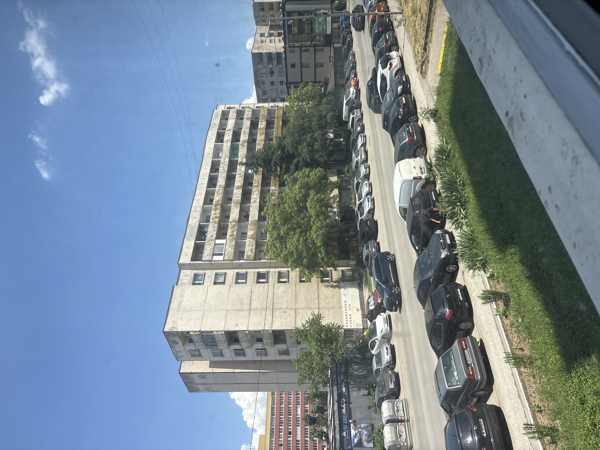 The image captures a street scene in Sofia, Bulgaria, on a bright, sunny day with a clear blue sky and scattered clouds. In the foreground, a row of parked cars lines the street, with a grassy area and some small bushes separating them from a concrete barrier. Beyond the cars, a multi-story residential building, characterized by its weathered, concrete facade and numerous balconies, dominates the middle ground. Trees with green foliage are situated in front of and around this building.
Further back, other buildings are visible, including a taller, red-and-white building to the left and several other apartment blocks. A commercial storefront with signage that includes "Avestauto" and possibly Bulgarian text is situated near the base of the large residential building. The overall impression is of a typical urban environment in Sofia.