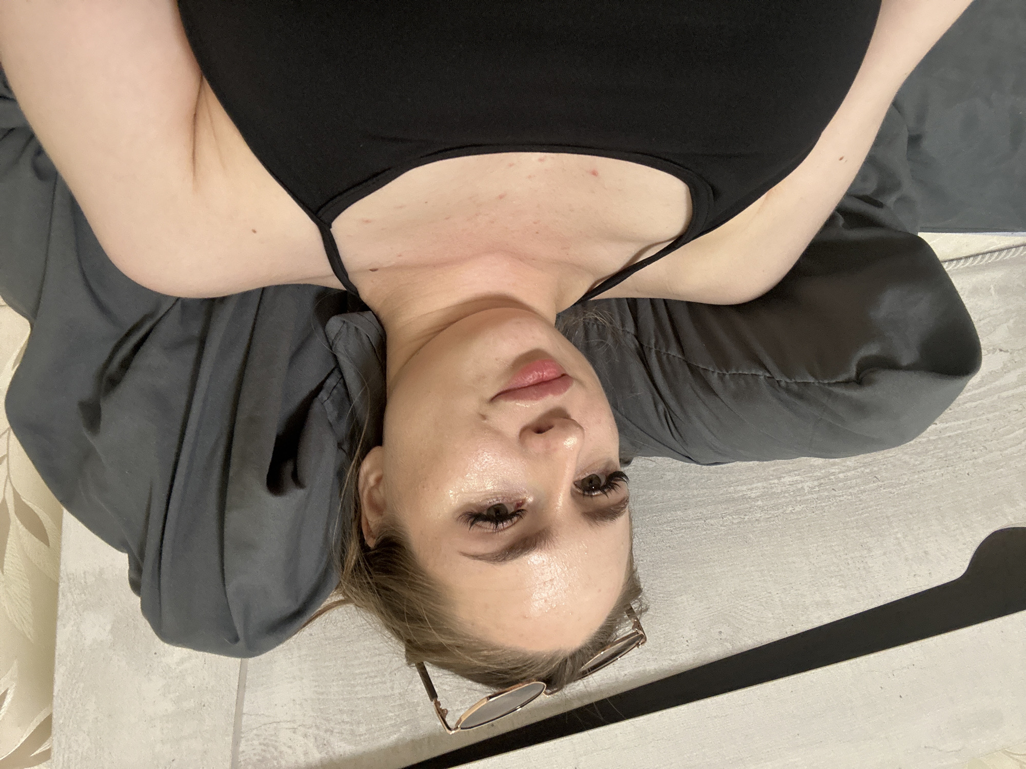 A female individual, appearing to be a young adult, is depicted in an indoor setting, likely a bedroom, in the city of Unknown, Unknown. She is lying on her back, supported by a dark gray pillow and bedding, and is looking directly into the camera. She has light brown hair, with a pair of sunglasses featuring a metallic frame resting on top of her head. Her skin has a noticeable sheen, and she has visible makeup, including defined eyelashes and lip color. Small reddish blemishes are present on her chest, and a singular mole is visible on her right upper arm. She is wearing a black tank top with thin shoulder straps. The background on the left features a light-colored, textured wall with a prominent horizontal dark stripe or shadow. On the right, a section of cream-colored wallpaper with a subtle leaf pattern is visible.