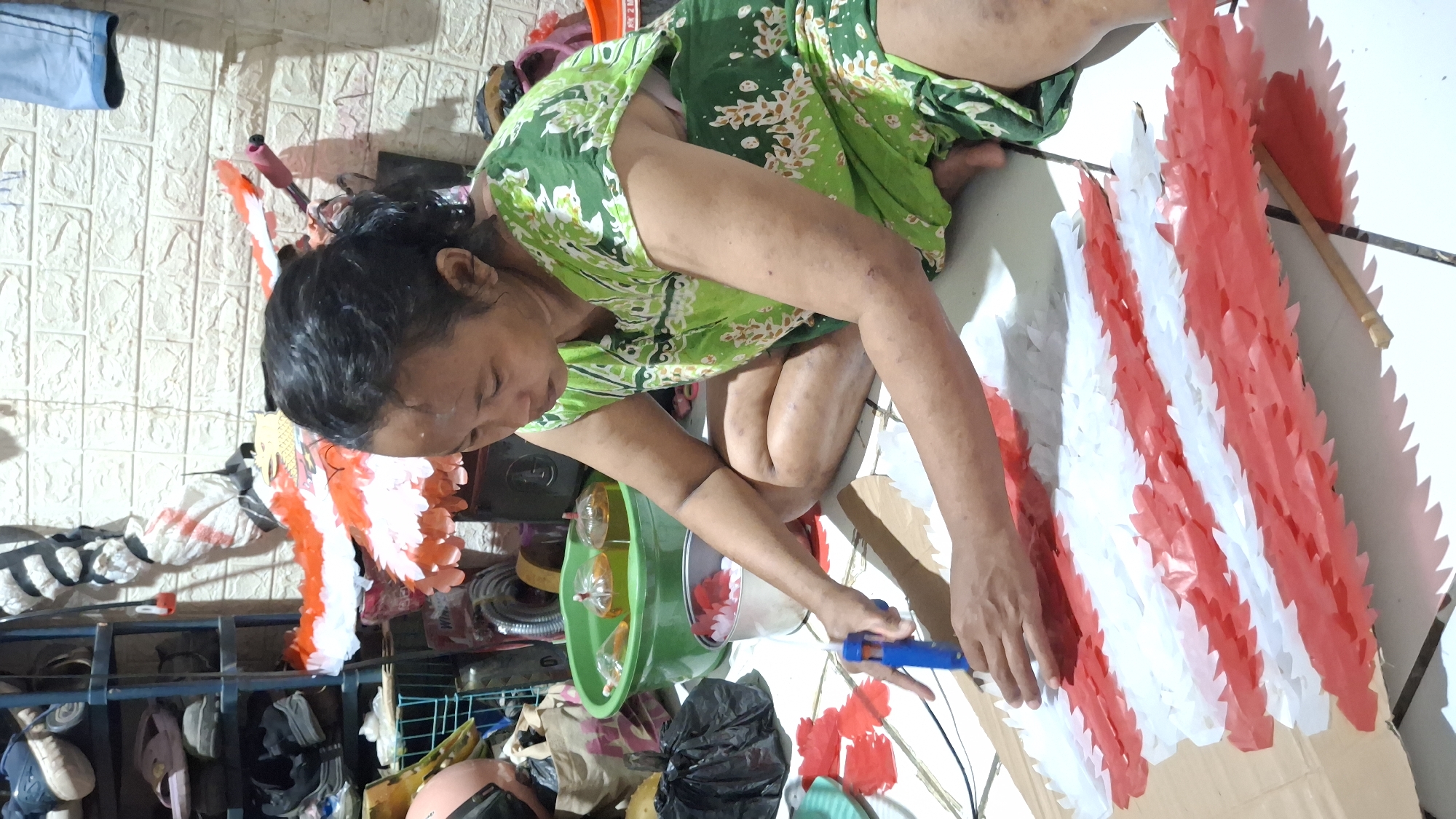 A woman sits on the floor of an indoor space, crafting with red and white paper. She is wearing a green and white floral sleeveless dress and has her dark hair pulled back. Her right hand holds a blue glue gun, which she is using to attach paper strips to a larger cardboard base. The paper strips are arranged in a pattern of alternating red and white, with a jagged edge. In the background, there is a shelving unit with various items, including shoes and baskets, and a wall with a textured, brick-like pattern. To her left, a green basin holds three glass containers filled with a yellowish liquid, possibly oil or candles. There are also black plastic bags and other craft supplies scattered around her. The flooring appears to be tiled.