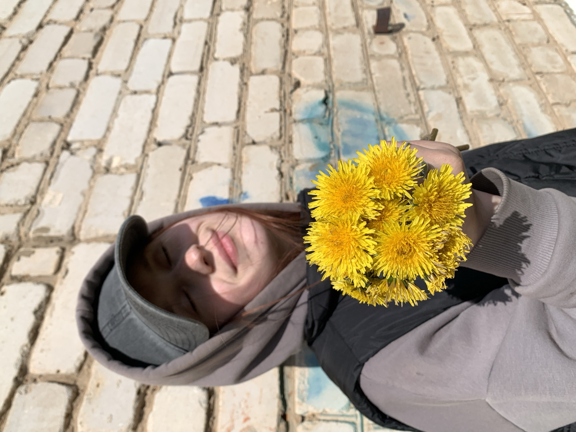 A high-angle, portrait-oriented photograph captures a young person resting outdoors in Фоки, Russia, holding a vibrant bouquet of yellow dandelions. The subject, seen from the chest up, is wearing a grey baseball cap, a grey hooded sweatshirt, and a black outer garment. Their eyes are closed, and a faint smile plays on their lips, suggesting a moment of relaxation or contentment. Bright sunlight illuminates their face and the dandelions.
The person's right hand holds a cluster of freshly picked dandelions, which are a prominent splash of bright yellow in the foreground. The background consists of a textured surface, likely a light-colored brick wall or ground, with visible mortar lines. Some faded blue and greenish paint marks are present on the brickwork behind the person's shoulder. Further up and to the right, a dark, indistinct, boot-shaped object or mark is visible on the wall. The bright, direct lighting indicates it is daytime and sunny. There is no visible text in the image.