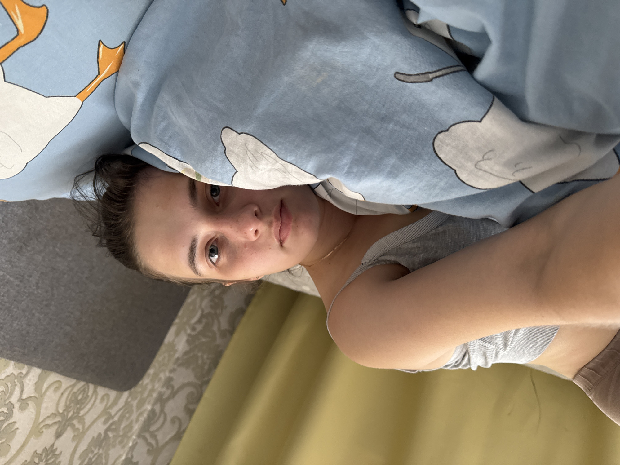 A woman is lying in bed, partially covered by a blue and white duvet. She is wearing a gray tank top. The background shows a patterned gray and white headboard and a yellow curtain. The location is likely Chaikovsky, Russia, but this is not visually verifiable from the image.