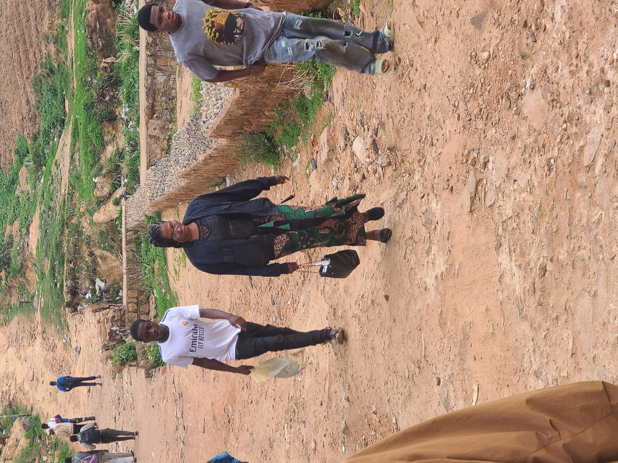 The image shows a dirt path in an outdoor setting in Unknown, Unknown. Three people are prominent in the foreground. One person, likely male, is wearing a grey t-shirt and ripped jeans. A second person is wearing a black jacket and patterned green dress, and the third person is wearing a white t-shirt and black pants, carrying a translucent yellow bag. In the background, additional people are walking along the same path. The area beside the path appears to be a mix of grassy areas and what may be a constructed retaining wall. The ground is a mix of dirt, rocks, and debris.