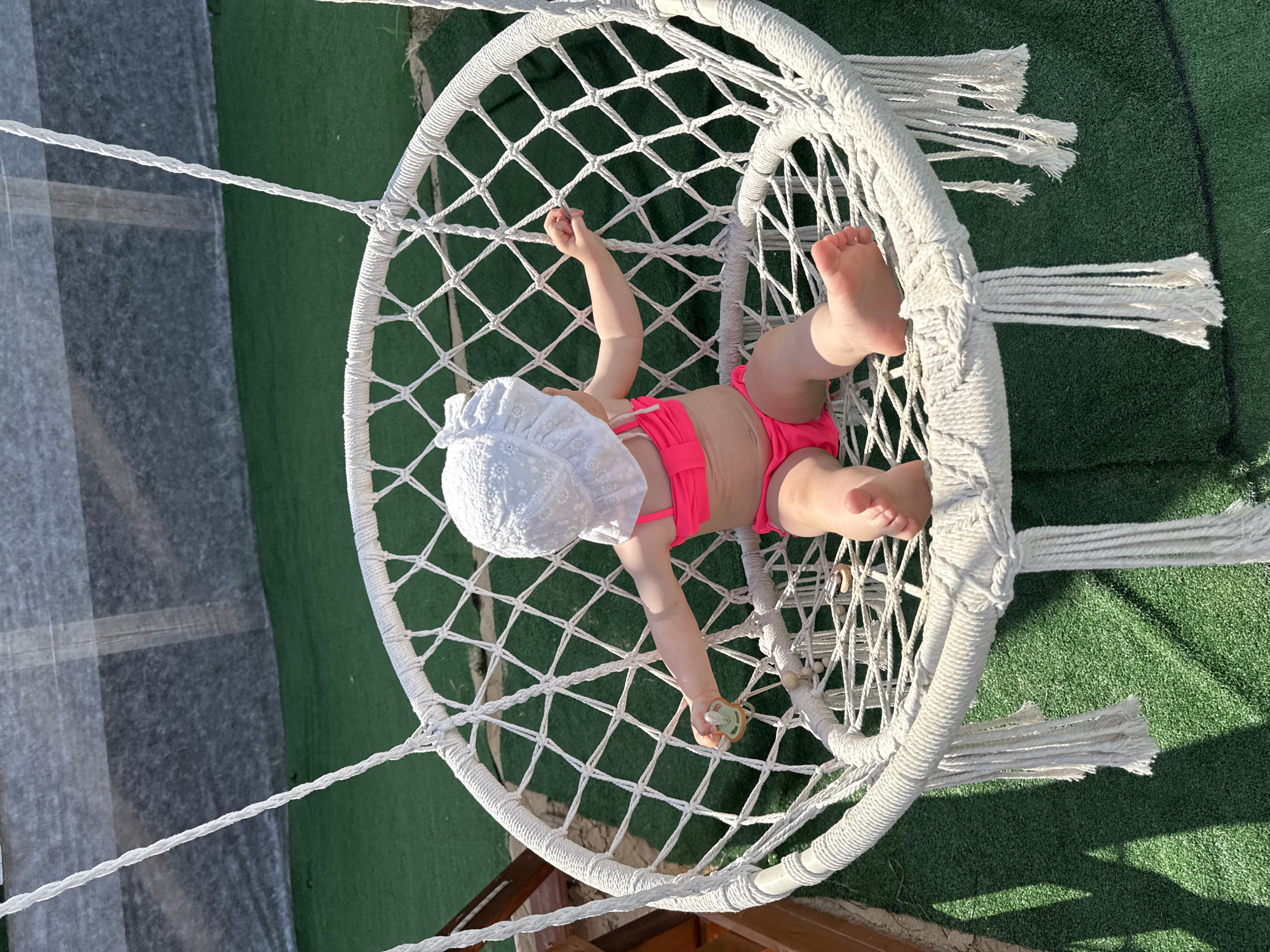 A baby is lying in a white macrame hammock chair. The baby is wearing a white sun hat and a pink bikini. The hammock is suspended from above by ropes and is hanging over a green, artificial turf surface. In the background, there is a blurred area that appears to be a gray tarp or sheet, possibly covering something. The scene is outdoors, and the bright sunlight suggests it is daytime, likely a warm day given the baby's attire. The overall impression is one of a relaxed, leisurely moment, perhaps at a backyard or resort setting. The baby's posture and the pacifier in its hand suggest a moment of rest or play. There is no visible text in the image.