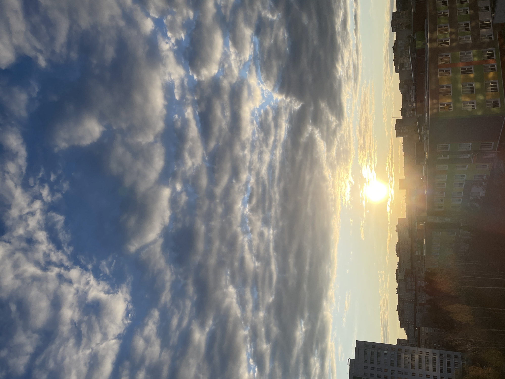 The image shows a view over the city of Izhevsk, Russia, at sunset. In the foreground, a cloudy sky is visible, with the sun shining through the clouds. In the background, there are several buildings, including residential apartment blocks. The buildings are of different heights and styles. The sky is partially blue. There are trees visible in the distance. The sun is low on the horizon, creating a warm glow over the city. The photo appears to have been taken from an elevated position, likely from a high-rise building.