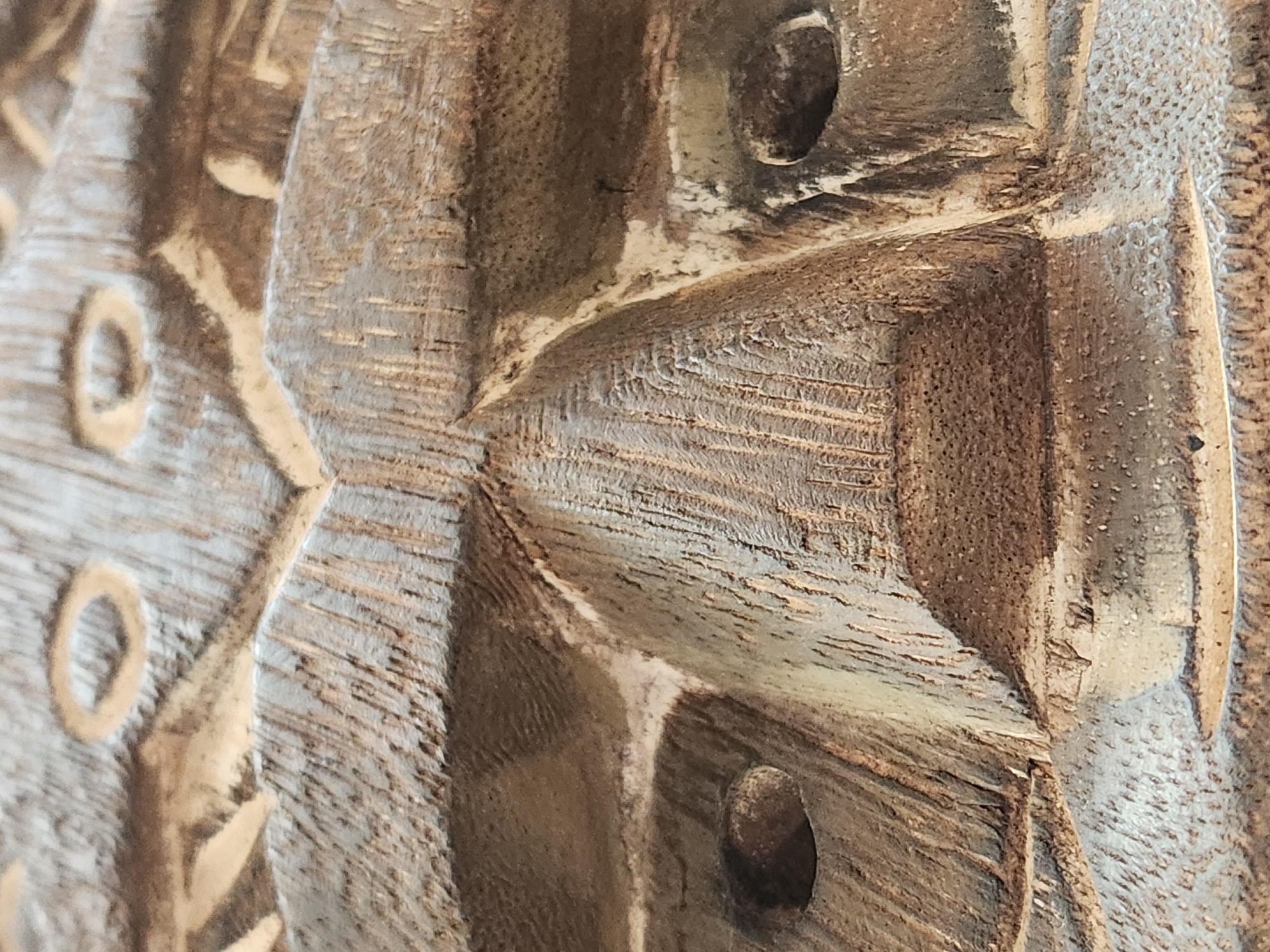 The image displays a close-up, angled view of a carved wooden mask. The mask features prominent, stylized eyes, a large, triangular nose with carved nostrils, and an indication of a mouth. The texture of the wood is visible, showing grain and rough carved surfaces. The overall color palette is earthy browns and grays. The image does not provide any information about the city of Maitland, United States of America, nor does it depict any people, actions, or interactions. It is purely a detailed shot of the carved artifact.