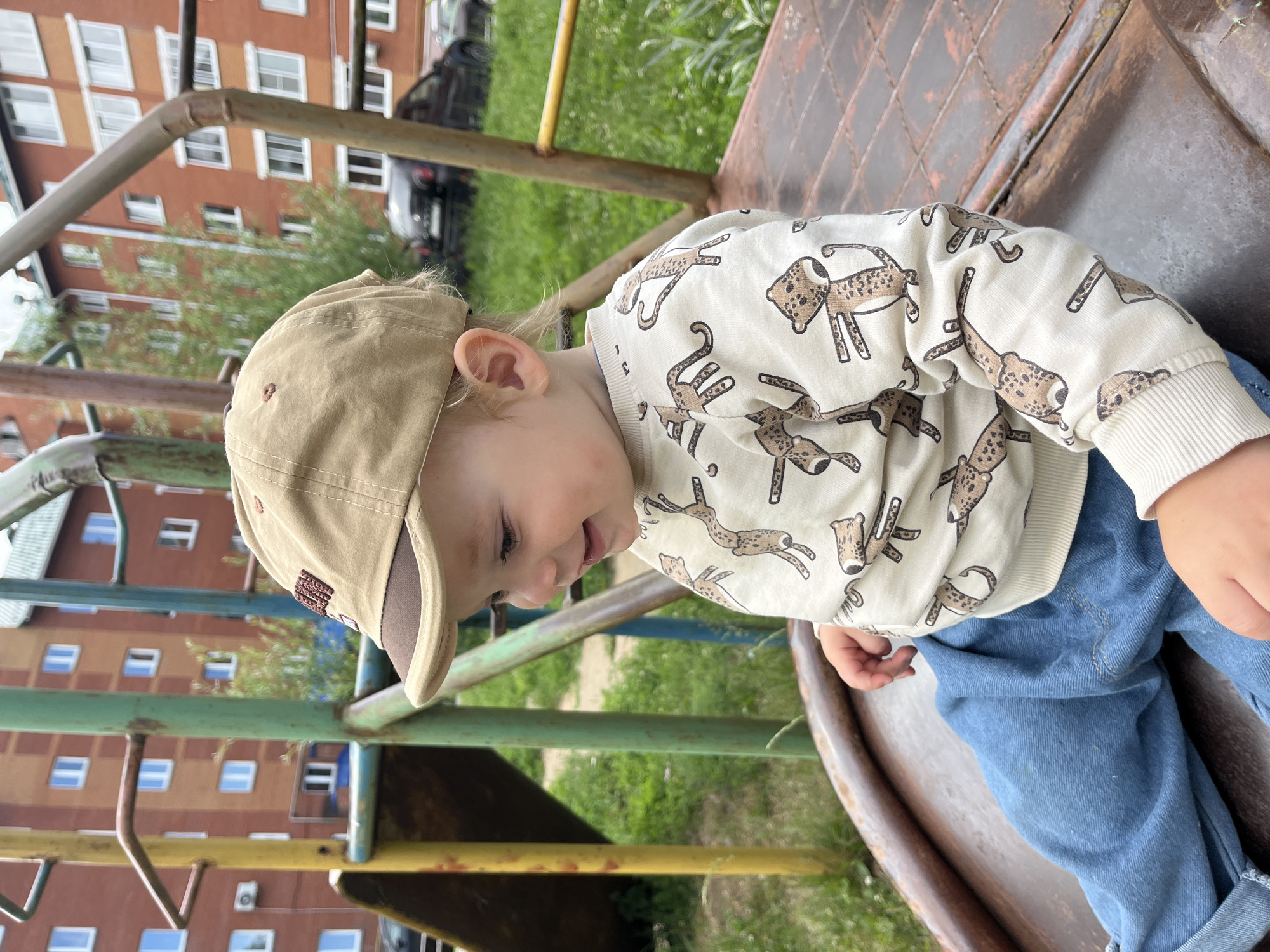 A young child wearing a beige baseball cap and a cream-colored sweatshirt with a leopard print sits on a rusty metal slide in a playground. The child is looking to the side with a slight smile. The playground equipment, painted in various colors including yellow, green, and blue, is visible in the background. Behind the playground, apartment buildings with orange brick facades and white windows are prominent. The ground is covered in green grass, and a few cars are parked in the distance. The scene appears to be set in a residential area in the city of Chaikovsky, Russia.