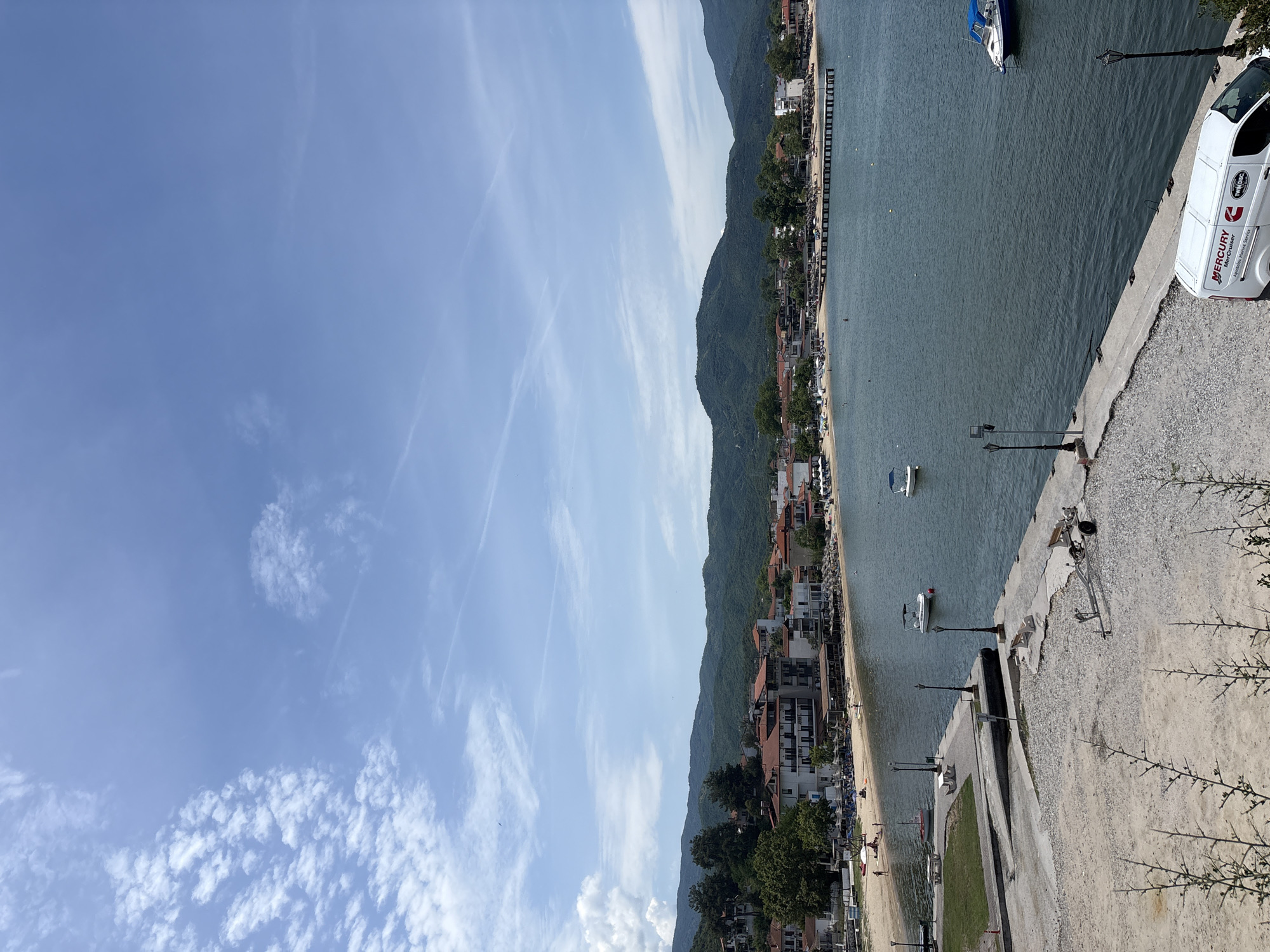 This image captures a scenic view of the coastal town of Olympiada, Greece. The foreground features a concrete embankment with a white van bearing "MERCURY MerCruiser" and "TWIN DISC" logos parked to the right. Further left on the embankment are some scattered pieces of lumber and a trailer. Beyond the embankment, a body of calm, dark blue water is visible, with several small boats and a larger white and blue boat anchored.
The middle ground reveals a sandy beach populated with sunbathers and umbrellas. Behind the beach, a row of buildings with red-tiled roofs and white facades lines the shore. These buildings appear to be hotels or restaurants. A long wooden pier extends from the beach into the water.
The background is dominated by lush green, rolling hills that rise steeply behind the town. The sky above is a bright blue with scattered white clouds, indicating fair weather. The overall impression is one of a peaceful seaside resort on a sunny day.