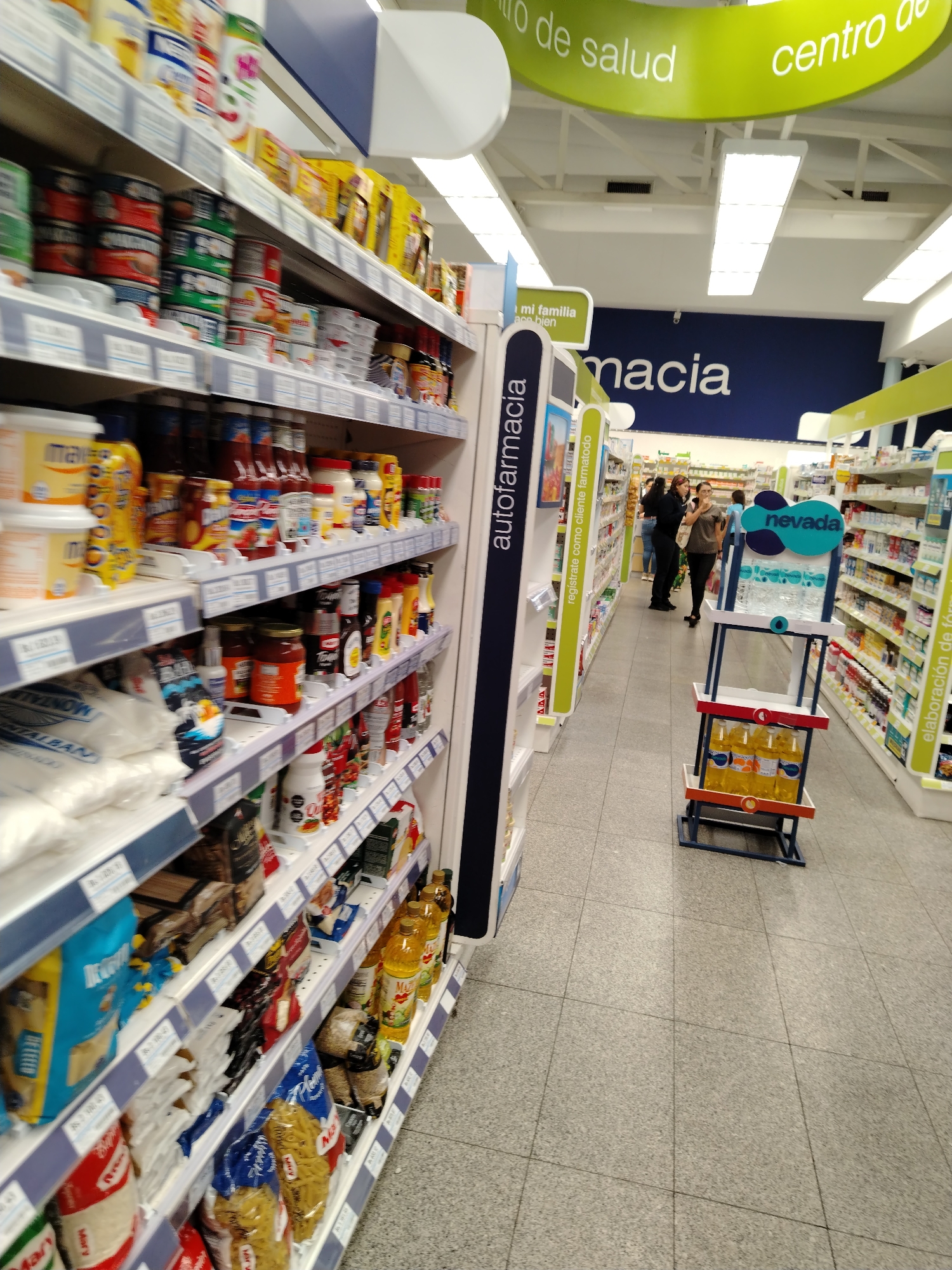 The image shows an aisle in a pharmacy, possibly a branch of Farmatodo in Tinaquillo, Venezuela, based on the signage. The shelves on the left are stocked with a variety of groceries, including canned goods, sauces, flour, pasta, and cooking oil. In the background, people are browsing the pharmacy section. There is a display rack with bottles of water and juice positioned in the center of the aisle. The lighting is bright and the floor is tiled. Signage indicates the presence of an "autofarmacia" and a "centro de salud."