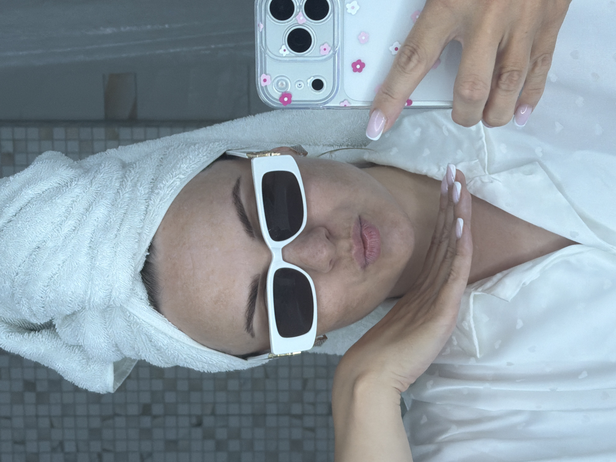 A woman lies on a tiled surface, possibly a bathroom floor, in what appears to be an indoor setting. She has a white towel wrapped around her head and is wearing large white sunglasses. She is dressed in a white satin shirt, and her fingernails are manicured with a French tip design. She is holding a phone in her hand with a clear case decorated with small pink flowers. The setting seems to be a private residence due to the personal nature of the scene. The location in the city of Votkinsk, Russia, cannot be determined from the image.