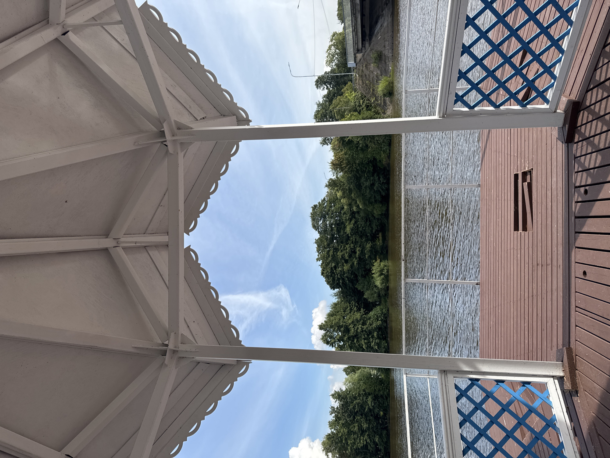 The image shows a view from inside a white gazebo, looking out at a body of water. The gazebo has a wooden deck floor and decorative blue and white lattice railings. Beyond the gazebo, a line of green trees borders the water, and a bridge with a road is visible in the distance. The sky is blue with some scattered white clouds. This scene is located at Dubki Park in Sestroretsk, Russia, a resort town on the Karelian Isthmus. There are no people or animals visible in the image.