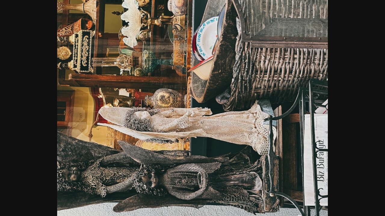 The image shows a display of antique-like objects, possibly in a shop or museum. The scene is vertically oriented and appears to be captured from a low angle, looking upwards.
In the upper portion of the image, there are shelves filled with various items. Some visible objects include ornate metalwork, glassware, and what appear to be decorative vases or jars. A white lace doily with a ruffled edge is prominent on the top shelf. Further to the right, a woven basket-like structure is visible, with metal framing and a mesh screen at the top. A stack of plates, one with a red and blue rim, is placed inside a dark, curved object.
Below, two figures, possibly dolls or mannequins, are laid out. The upper figure is dressed in a cream-colored, lacy shroud. Its head is visible and appears to be made of pale material, with curly hair made of thread. Below this figure, another figure is similarly draped, but this one is covered in darker, more coarse material, also with lacy details and a visible head with similar curly hair. Both figures are positioned horizontally.
To the right of these figures, a metal stand supports a tall, white rectangular object with text on it. The text is in a foreign language, but "Biz Bura" is partially legible.
The overall impression is of a cluttered yet curated collection of vintage or antique items, with a somber or gothic aesthetic suggested by the draped figures. The lighting is somewhat dim, creating shadows and highlighting the textures of the objects. The background is a mix of wooden shelves, glass reflections, and dark areas.