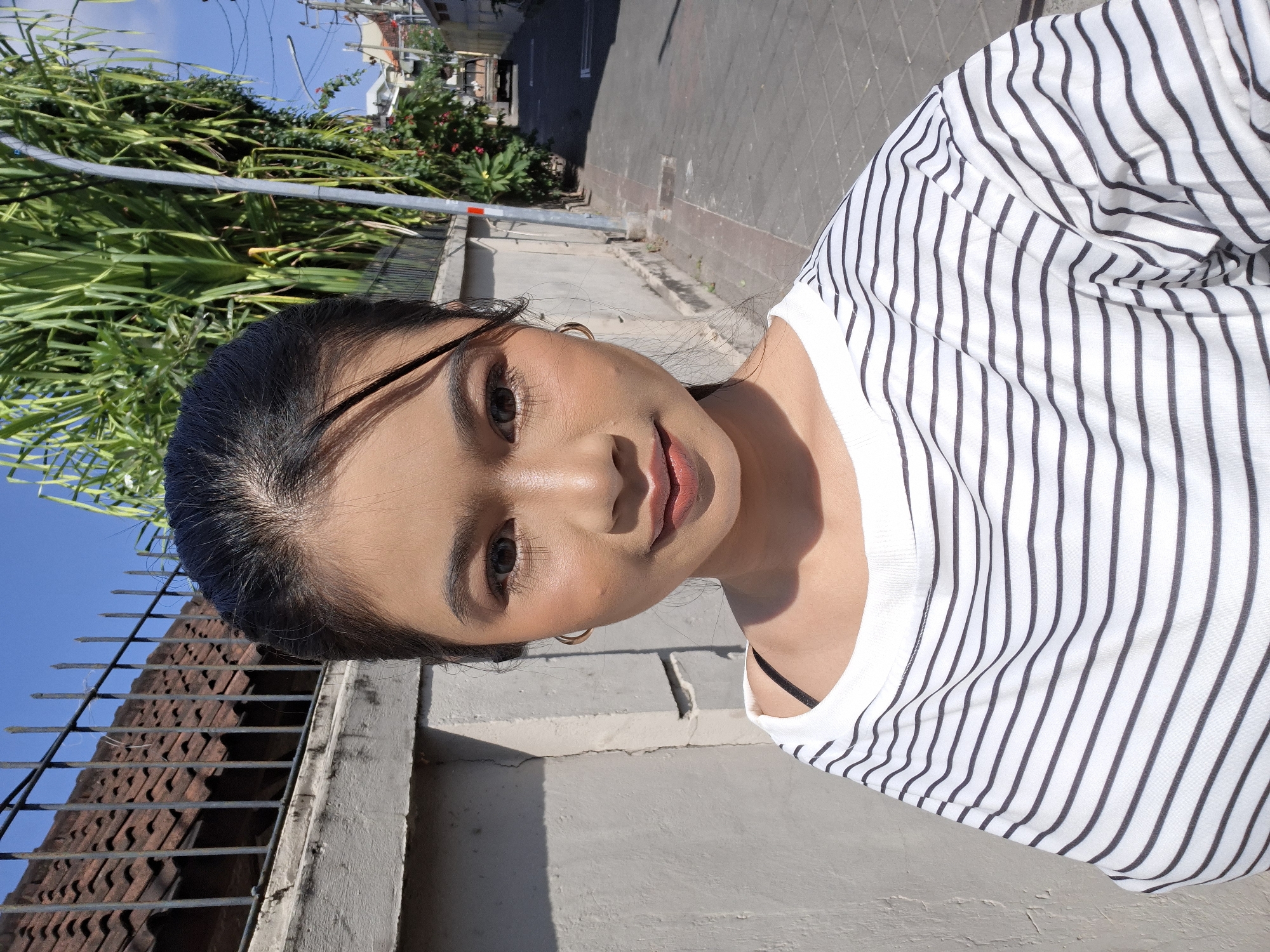 A young woman with dark hair pulled back in a ponytail and wearing makeup poses for a selfie in Kuta, Indonesia. She is wearing a white t-shirt with black stripes and a black bra strap is visible. The background shows a paved street, a concrete wall, a metal fence with a tiled roof behind it, and lush green foliage with a street lamp. The sun is shining brightly, casting shadows. In the distance, a vehicle can be seen on the street.