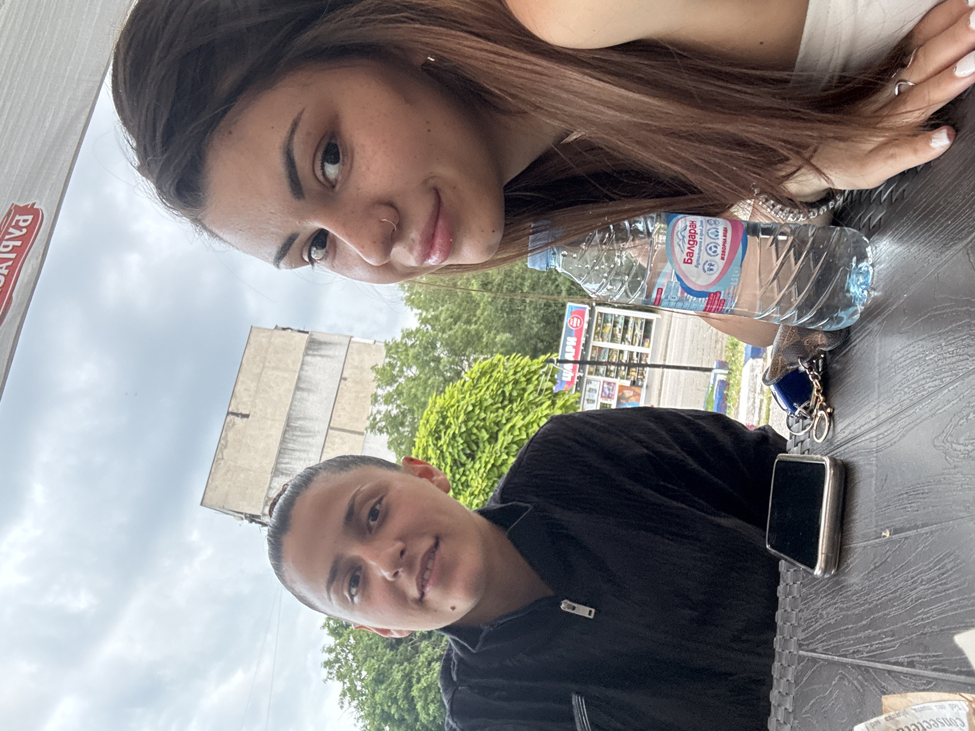 Two young adult females are seated outdoors at a dark, textured table, likely a cafe, under an awning with inverse red Cyrillic lettering. The woman on the left, with dark hair pulled back, wears a black collared top with a zipper and looks towards the camera with a slight smile. The woman on the right, partially visible with long brown hair, wears a light-colored top, has a nose ring, and displays multiple rings and white nail polish on her right hand as she smiles towards the camera.
On the table between them are a silver-colored smartphone, a set of keys with a blue fob, and a 1.5-liter plastic bottle of "Балдаран" (Baldaran) spring water, featuring prominent Cyrillic labeling. The background includes green trees, a light-colored multi-story building, and a distant commercial kiosk with red and blue Cyrillic signage, indicating an urban setting. The sky is overcast. This scene is located in the city of Sofia, Bulgaria.