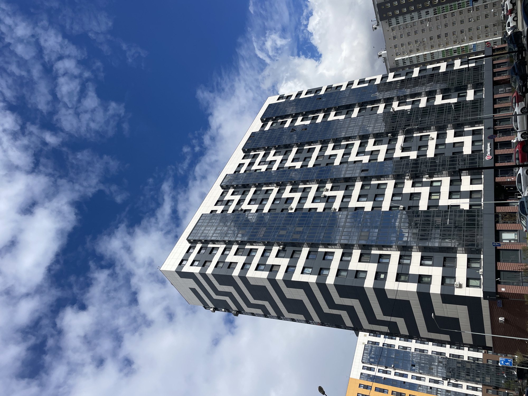 The image displays a modern, tall building with a distinctive geometric facade set against a bright blue sky with scattered white clouds. The building, viewed from a low-angle perspective, features a striking black, white, and gray zigzag pattern on its exterior walls, which incorporates numerous windows. Upper sections of the building are clad in dark, reflective glass panels that mirror the sky and clouds.
In the background, a more traditional, gray high-rise building with a grid of windows is visible. At street level, a row of parked cars lines a road adjacent to the main building. The clear blue sky and bright lighting indicate a sunny daytime.
Visible text and signs provide contextual clues:
- A prominent red circular sign with a white "5" and the Cyrillic word "Пятерочка" (Pyaterochka) is affixed to the building, indicating a retail store, likely a supermarket.
- A blue parking sign with a white "P" is visible.
- A blue pedestrian crossing sign with white figures is also present.
- A street sign reading "ул. Г. Баруди, 14" (ul. G. Barudi, 14), which translates to G. Barudi Street, 14, is partially visible, confirming the location in an urban area, consistent with Kazan, Russia.
The scene captures a static urban landscape, highlighting contemporary architecture and everyday city elements under a clear sky.