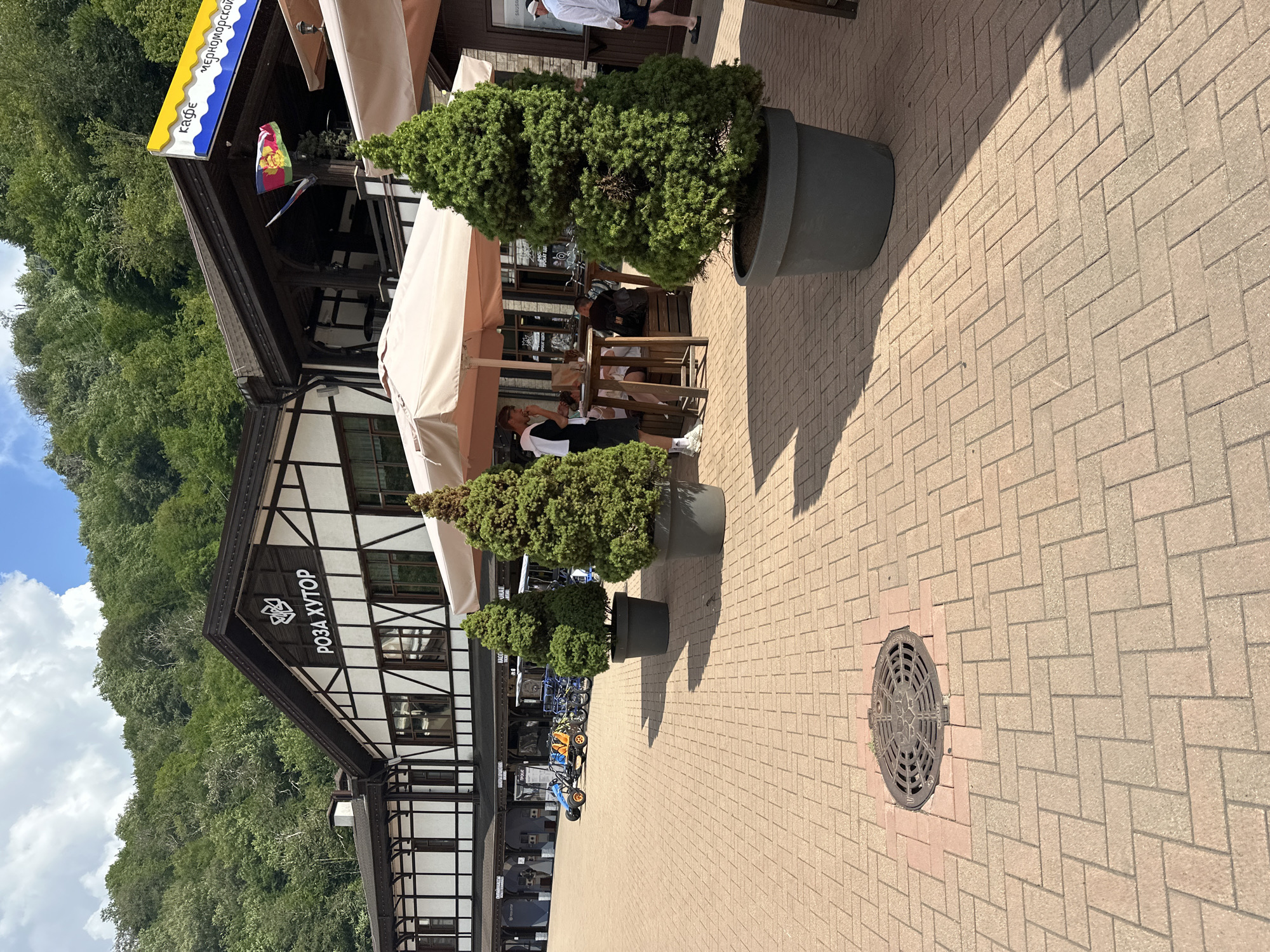 In the city of Estosadok, Russia, a bustling outdoor seating area is depicted in front of a building with a distinctive Tudor-style facade, featuring dark wooden beams against white stucco. The building is labeled with the Cyrillic text "РОЗА ХУТОР", which translates to "Rosa Khutor". Several people are present; some are seated at wooden tables beneath beige umbrellas, engaged in conversation or eating. One man, wearing a black and white top and shorts, stands by a table. Another man in a white shirt and shorts walks past in the background. The area is paved with light brown interlocking bricks, with a manhole cover visible on the ground in the foreground. Several large, dark grey planters contain conical evergreen trees, adding a touch of greenery to the scene. In the mid-ground, a small blue and orange ATV-like vehicle and several bicycles are parked. Flags, including what appears to be the flag of Adygea, are displayed on poles attached to the building. The background shows a lush, green forested mountainside under a bright sky with scattered clouds.