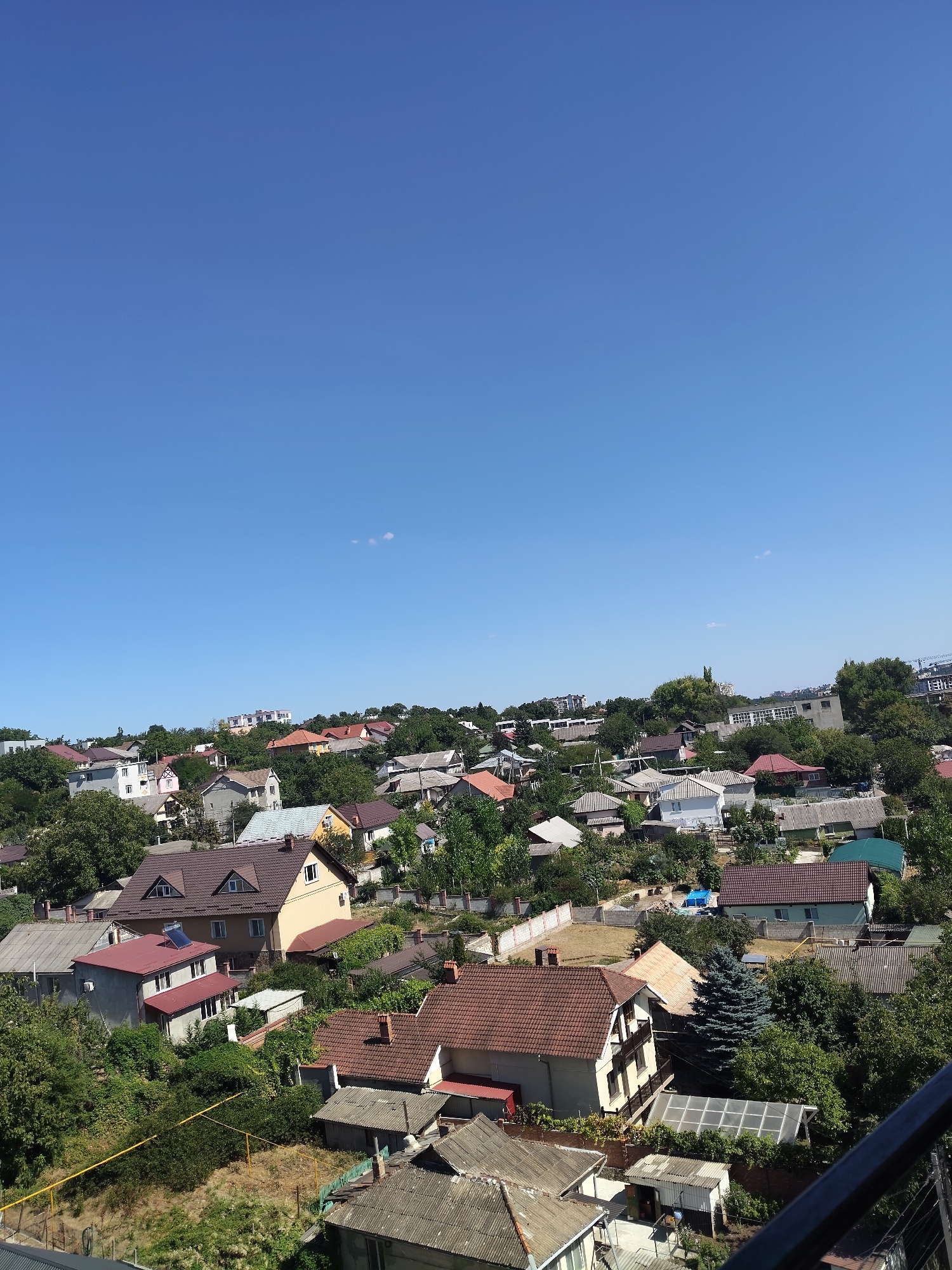 The image is an aerial view of a residential area in Durlești, Moldova, on a clear day. The scene is dominated by numerous houses with terracotta tile roofs, interspersed with dense green trees. The houses vary in size and architectural style, with some appearing to be older, single-story structures and others more modern, multi-story buildings. In the foreground, several houses are visible, including one with a prominent brown roof and dormer windows, and another with a red roof and solar panels. Further into the background, more houses and trees spread across a gently sloping landscape. The sky is a vibrant, cloudless blue, occupying a significant portion of the upper frame. The overall impression is of a typical suburban or semi-rural neighborhood. No people are visible in the image.
