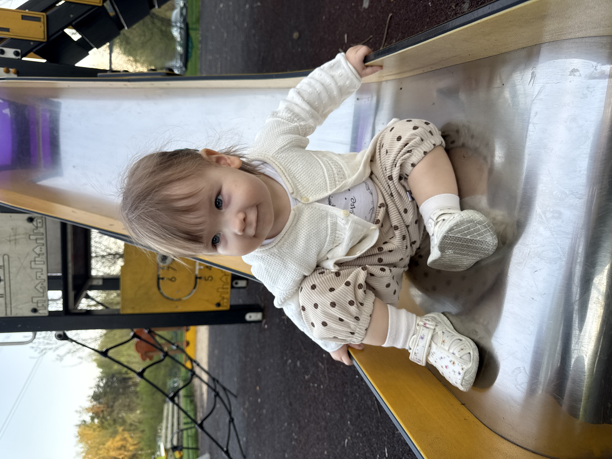 A young child is sitting on a playground slide, inside the city of Moscow, Russia. The child has light skin and hair, blue eyes, and is wearing a white cardigan, a white shirt, and polka-dotted pants. The slide is made of metal with a yellow frame. Other playground equipment is visible in the background, including a climbing net and a yellow and black structure with numbers on it. The ground is dark gray. Trees can be seen in the distance. The lighting is soft and diffused.