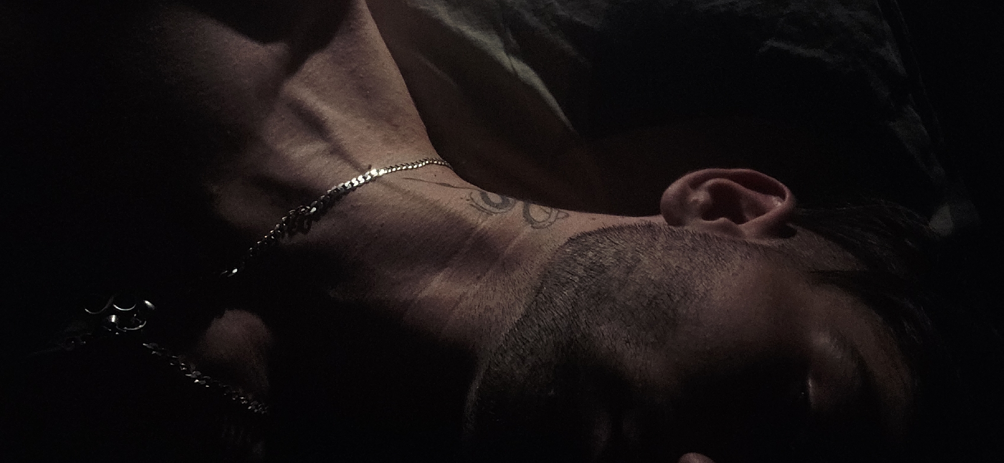 A close-up, dimly lit, overhead shot captures a portion of a man's upper body, focusing on his neck, shoulder, and the side of his face. The man has short, dark hair and a stubbly beard. He is wearing a silver chain necklace with a circular pendant. A tattoo of intertwined symbols is visible on his neck. The lighting creates strong shadows, obscuring much of his face and the background, which appears to be bedding. The scene is set in Kostenets, Bulgaria.