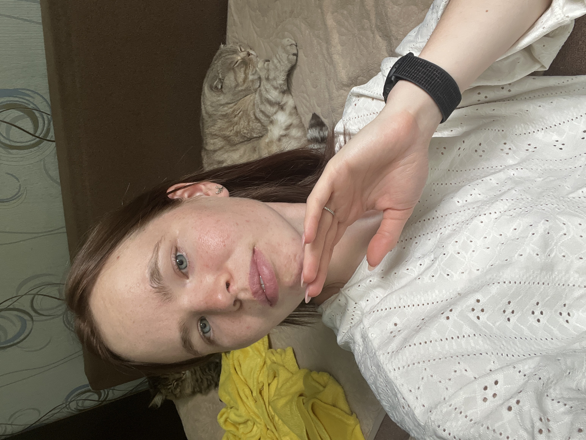 A woman lies on a brown sofa next to a cat. She has light skin, light blue eyes, and light brown hair. She is wearing a white shirt with eyelet fabric and a black smart watch on her left wrist. She is wearing a ring on her left hand. To her left is a yellow towel. The location is not discernible from the image.