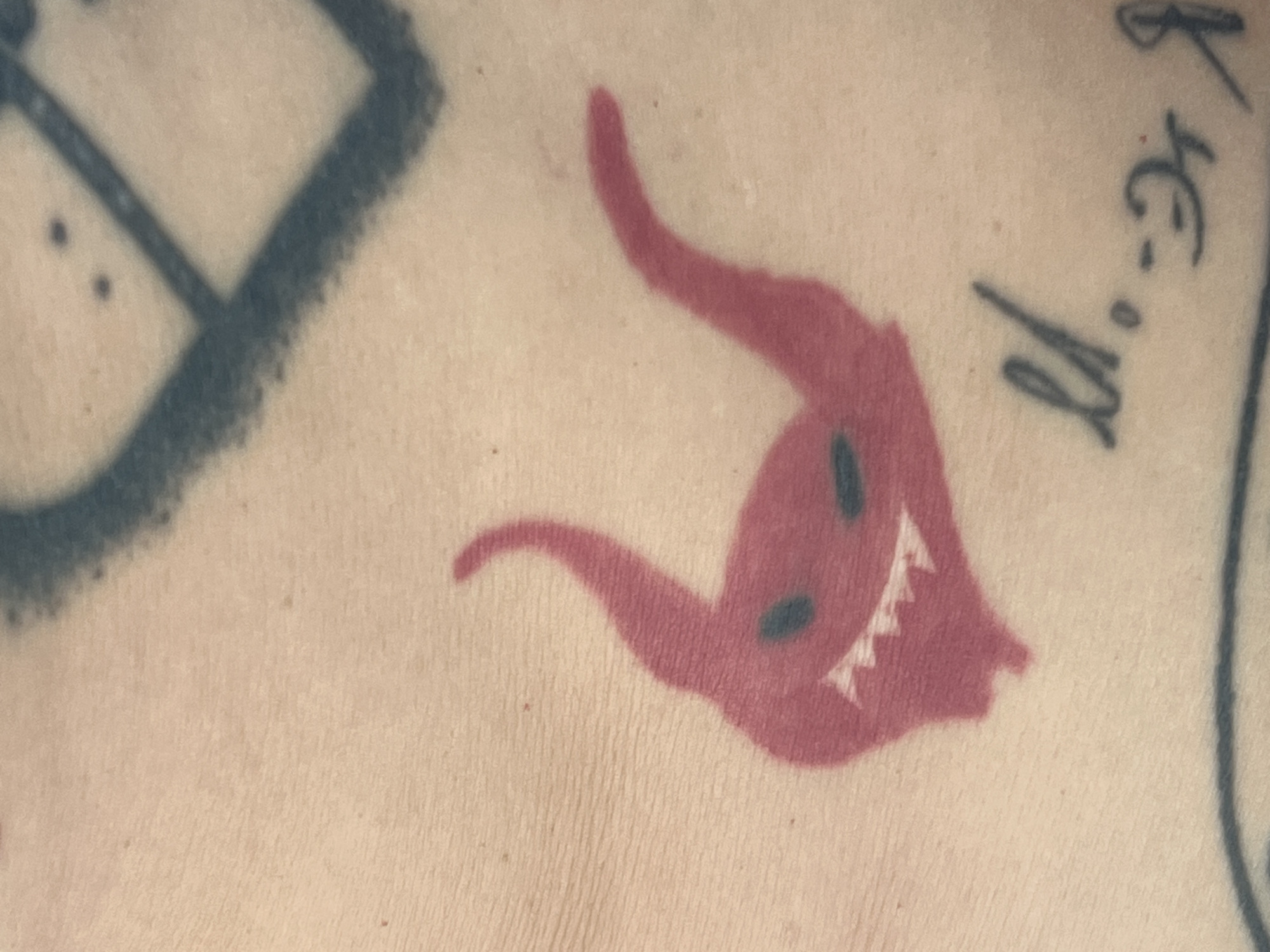 A close-up, top-down view of a tattoo on human skin is shown. The tattoo is of a small, red, cartoonish devil with two horns, two black eyes, a jagged white smile, and a pointed chin. The skin shows faint texture and a few small moles. In the background, a blurry, dark blue tattoo of a geometric shape is visible, as well as some script-like lettering. The lighting is even, with no harsh shadows. The location is unknown.