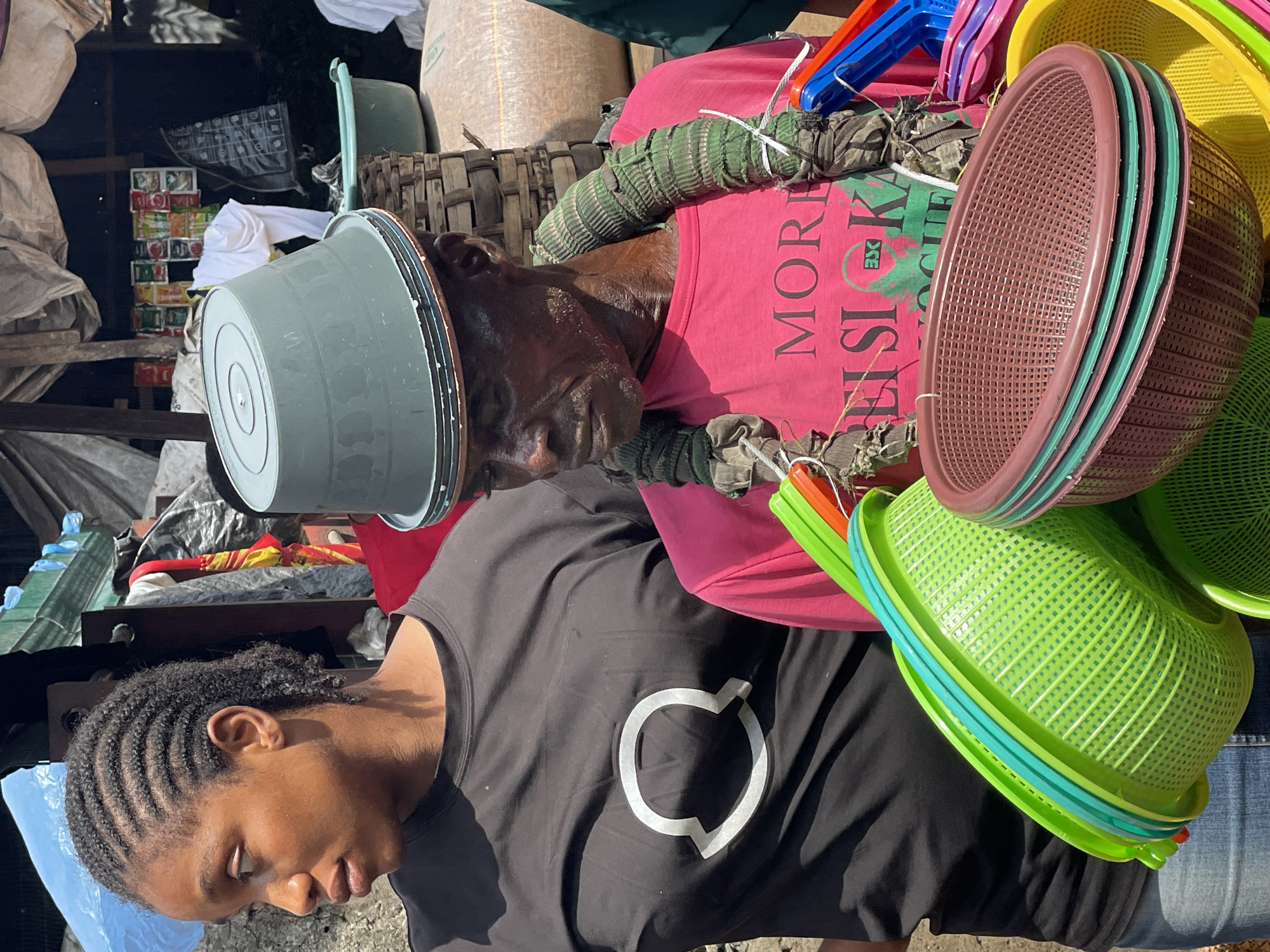This outdoor market scene in the city of Unknown, Unknown, features two individuals and various merchandise under bright daylight.
On the left, a young woman with dark skin and cornrow braids is shown in profile, looking to her left. She wears a black short-sleeved t-shirt with a white outline of the Moncler logo on the chest, and blue denim jeans.
On the right, an older man with dark skin has a grey plastic basin inverted on his head as a hat. He wears a bright pink t-shirt with "MORE" and "EISLOKA 3X" text printed in green and black. Two thick, camouflage-patterned fabric tubes, secured with white string, are draped around his neck and over his shoulders. He looks downward and slightly to his left.
In the foreground, a collection of new plastic baskets and colanders is prominently displayed. These include bright green items stacked on the left, and a stack of maroon-brown oval baskets with teal rims in the center-right. Additional yellow, pink, blue, and orange plastic items are visible behind these stacks.
The background shows elements of an informal market stall. Behind the man, a woven basket and a light green plastic basin are visible, along with a large woven sack. Further back, colorful packaged goods are displayed on a dark surface, beside a black patterned bag and a red and yellow umbrella. Overhead, various tarpaulins (light blue, grey, white, tan) cover makeshift structures.