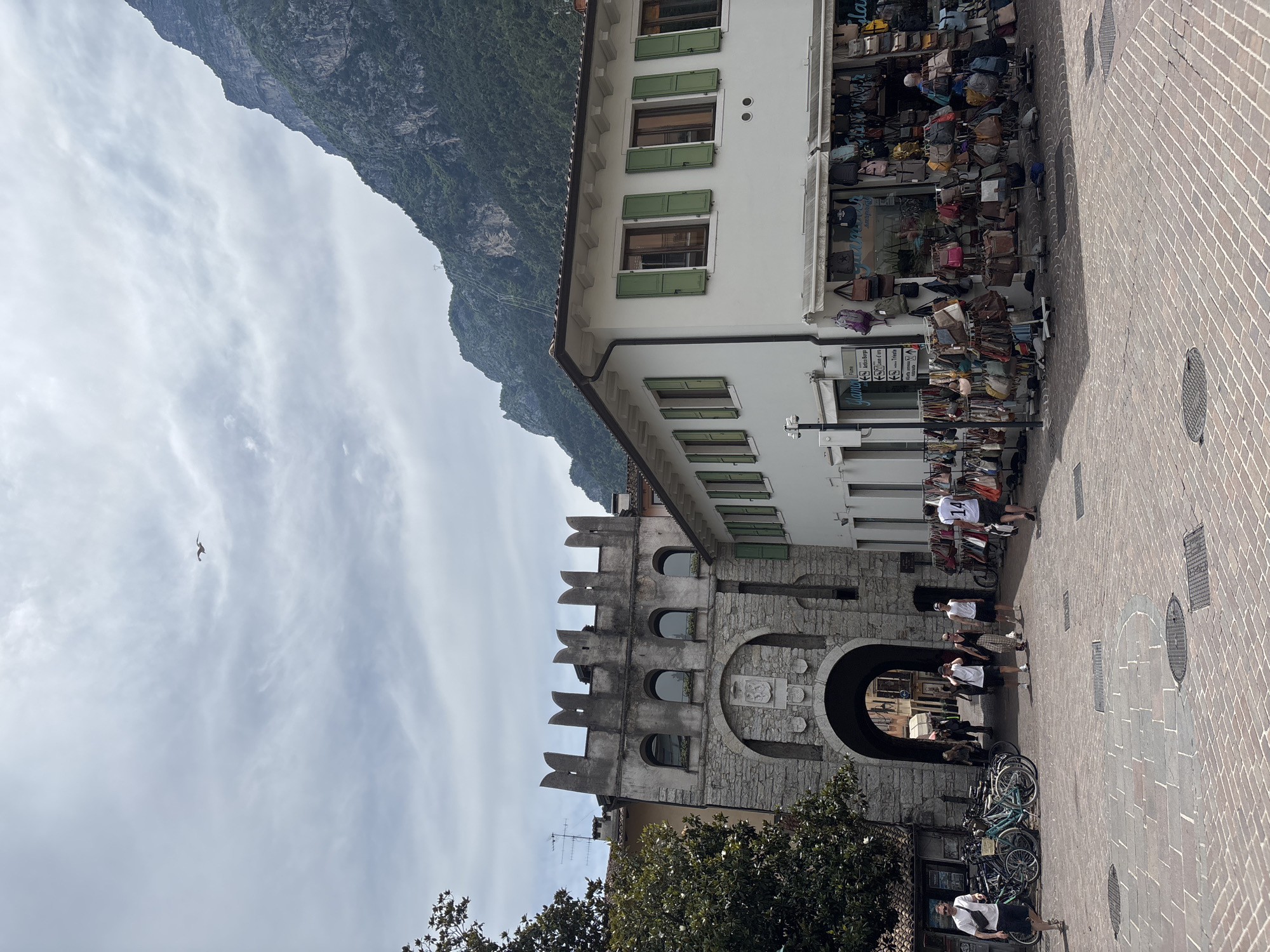In Riva del Garda, Italy, a bustling scene unfolds in a cobblestone piazza. An ancient stone gatehouse, adorned with arched windows and a coat of arms, stands prominently to the left. Beyond the gate, narrow streets hint at further exploration. To the right, a white building with green shutters houses a shop overflowing with handbags displayed on racks outside. A few people are visible in the square: a man with a white fanny pack walks to the left, while a group of four individuals, including a person walking a small dog, stroll towards the gatehouse. Behind them, another person in a white shirt with the number "14" on the back walks away from the camera. In the background, a lush green mountain looms under a cloudy sky, with a single bird soaring overhead. The overall impression is a vibrant snapshot of a European town, blending historical architecture with modern commerce and the natural beauty of its surroundings.