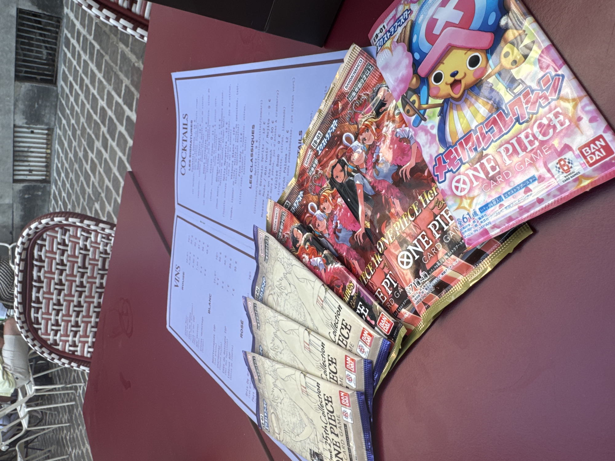 A table at a cafe in Lille, France, is covered by a red tablecloth. On the table are several packets of One Piece card games, arranged in a fan shape. A menu is also visible on the table, displaying a list of VINS (wines), COCKTAILS, and LES CLASSIQUES (classics). The cafe appears to be outdoors, with chairs and part of a building visible in the background. One person, wearing a light-colored shirt and shorts, is partially visible seated in a chair in the background. The packets of card games are colorful, with illustrations of characters from the One Piece anime series. The menu is in French. The packets appear to be from the "25th Collection". The atmosphere suggests a relaxed setting, possibly a cafe or restaurant.