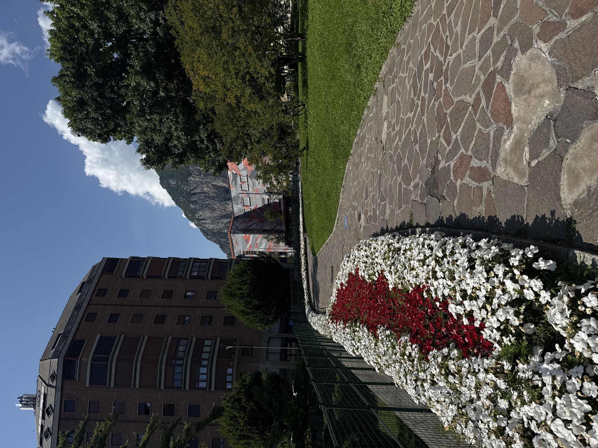A paved walkway curves through a park with a large flowerbed of red and white flowers along its edge. To the left of the walkway is a dark green metal fence and a multi-story brick building with numerous windows. In the background, a large, modern building with an orange and gray abstract pattern is visible, as are trees and a steep, rocky mountain under a blue sky with scattered white clouds. A lamppost stands near the brick building, and a cell tower or antenna array is on the roof of the brick building.