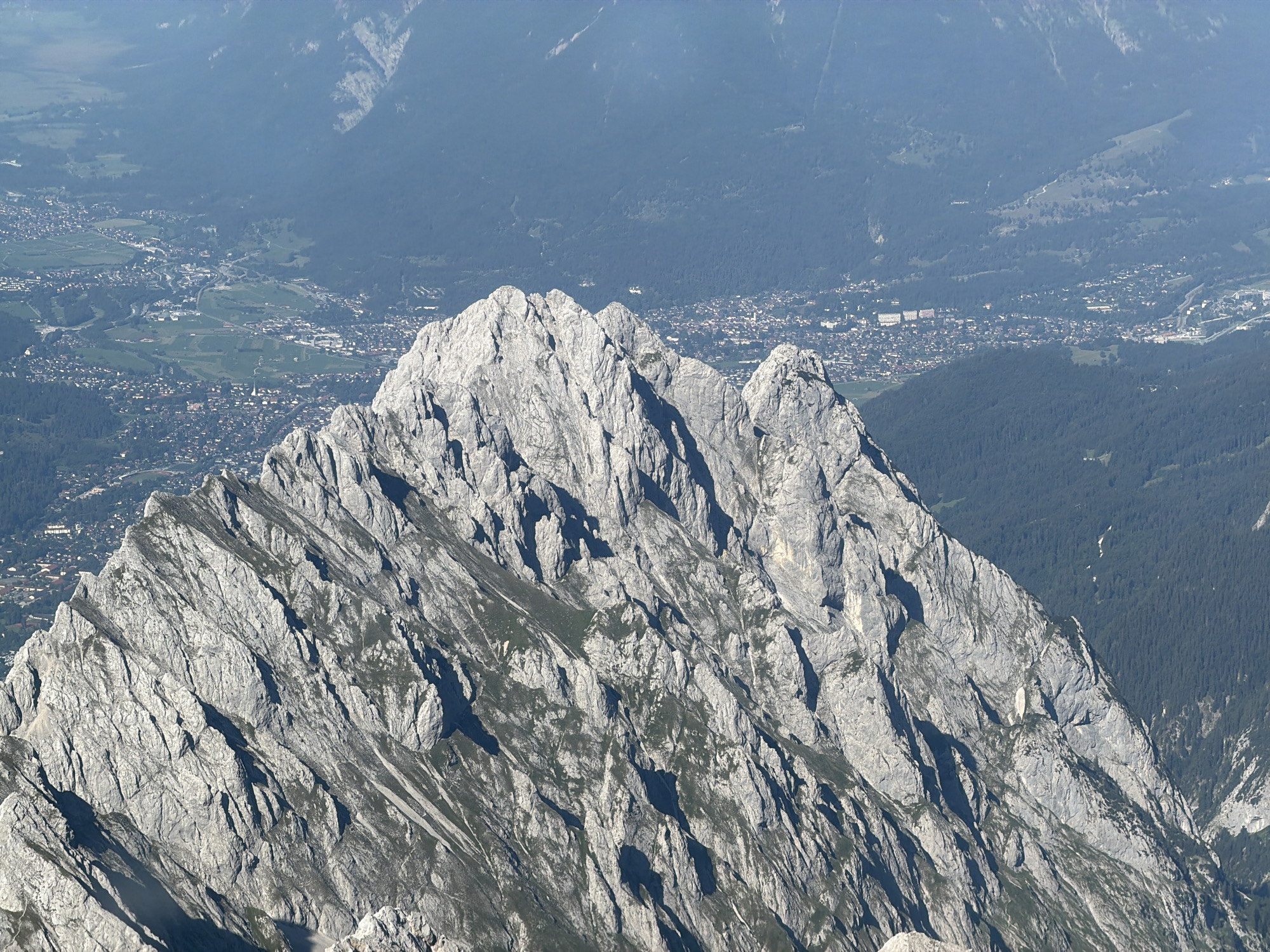 The image shows a panoramic view from a high mountain peak overlooking the city of Garmisch-Partenkirchen, Germany. In the foreground, a rugged, grey mountain range dominates the scene, characterized by sharp peaks, deep crevices, and sparse vegetation clinging to its slopes. The sunlight casts dramatic shadows across the rocky terrain. In the mid-ground and background, the city of Garmisch-Partenkirchen is visible, spread across a valley floor. The urban landscape is a tapestry of buildings, roads, and green spaces, nestled amongst forested mountains. The distant mountains appear hazy, fading into a soft blue in the upper portion of the image, suggesting a clear but distant view. There are no people or specific actions depicted in the image. The overall environment is one of vast natural beauty and the presence of a human settlement within a mountainous region.