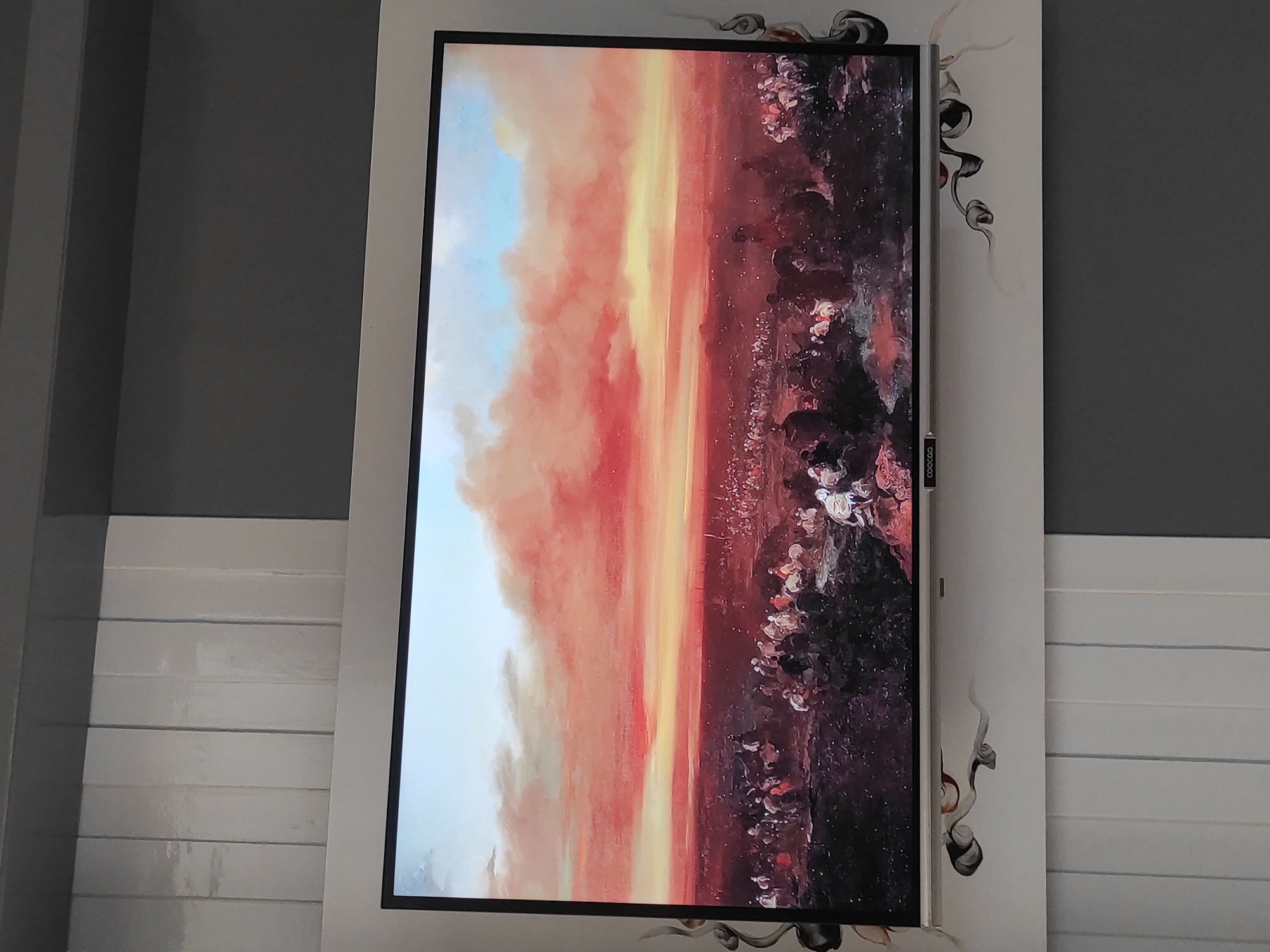 The image shows a large television screen displaying a painting of a historical battle scene. The painting depicts a vast landscape with a dramatic, fiery sunset. In the foreground, numerous figures on horseback and on foot are engaged in what appears to be a conflict. The figures are rendered in a realistic style, with attention to detail in their clothing and armor. The setting is arid and rocky, with sparse vegetation. The sky is filled with vivid hues of orange, red, and yellow, indicative of a sunset or sunrise. The television itself is a flat-screen model with a thin black bezel, mounted on a wall. The wall is decorated with a patterned wallpaper. The lower portion of the image shows blinds or curtains, suggesting the television is in a domestic setting. There is no discernible connection to the city of Sumedang, Indonesia, as the scene is a generic historical painting.