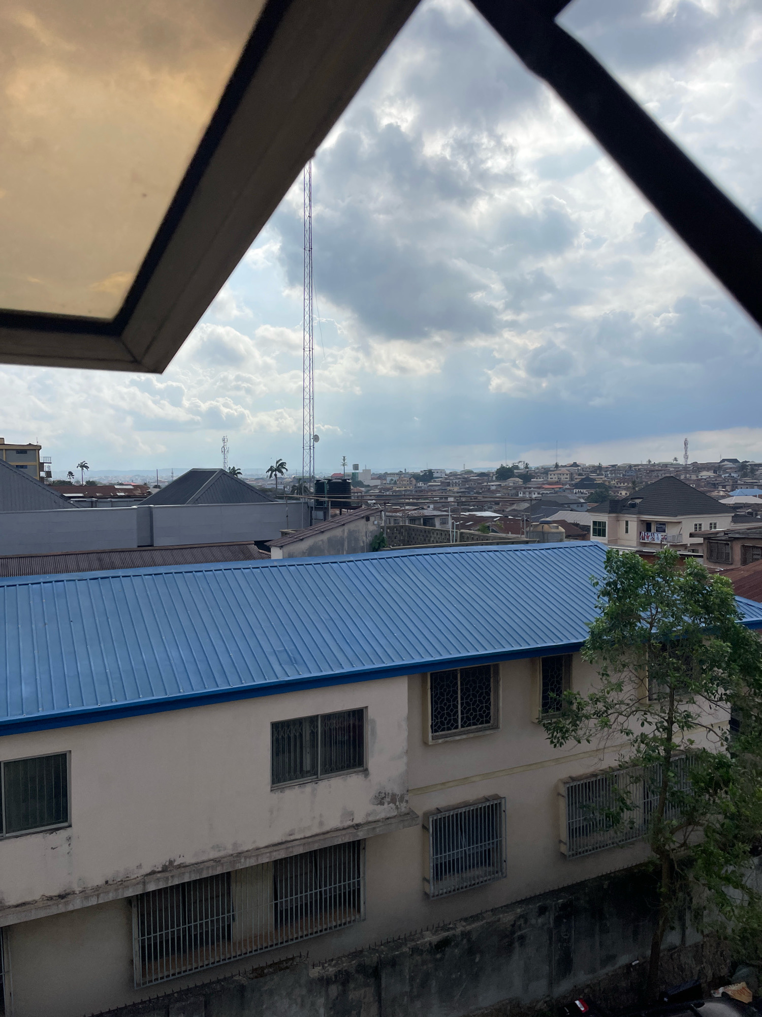 The image captures a view of the cityscape of Ibadan, Nigeria, from an elevated position, likely a building. In the foreground, a section of a window frame with a yellow-tinted glass occupies the top left of the frame. Below this, the prominent feature is the blue corrugated metal roof of a building. The building itself has a light-colored facade and several windows, many of which are barred. A tree with green leaves stands to the right of this building.
Beyond the immediate foreground, the landscape unfolds into a dense urban environment. Numerous buildings with various roof types, including metal and tiled roofs, stretch towards the horizon. The city appears somewhat hilly in the distance. Several telecommunication towers are visible throughout the cityscape, indicating a developed communication infrastructure. The sky is overcast with dramatic, grey clouds, suggesting it might be a cloudy day or that rain is impending. No people are visible in the scene. The overall impression is of a typical urban environment in Ibadan, characterized by its density of buildings and infrastructure, set against a cloudy sky.
