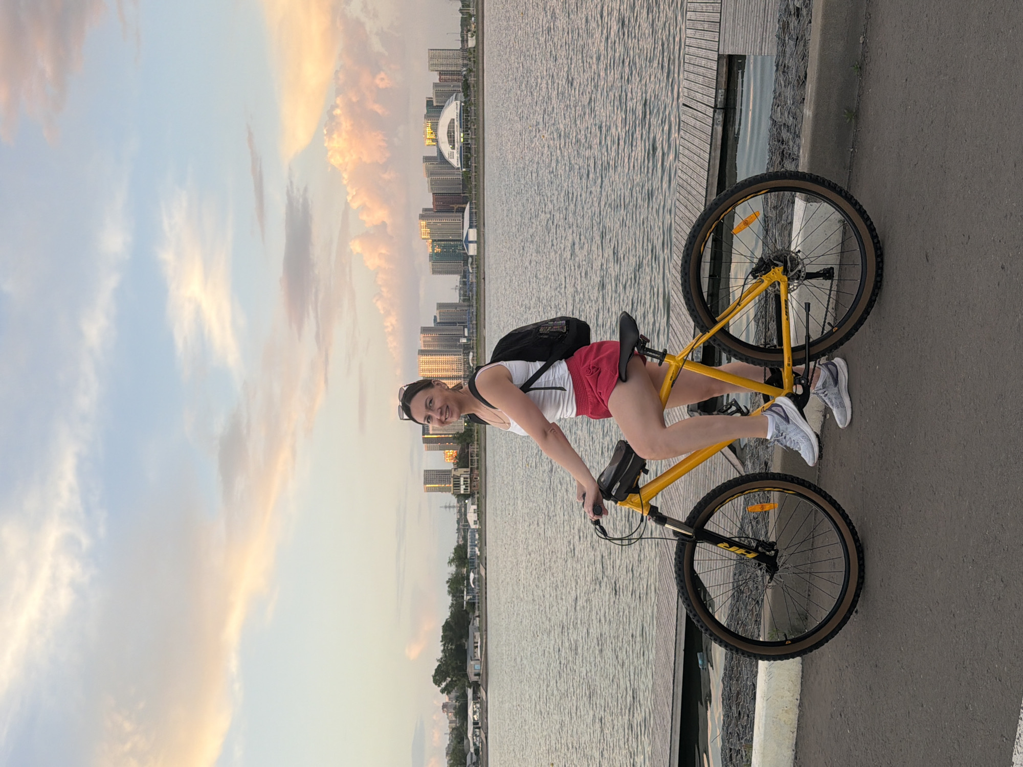 A woman is posing on a yellow bicycle next to a body of water. She is wearing a white tank top and red shorts with gray sneakers and has a black backpack on her back. The backdrop consists of a body of water, a wooden dock, a paved pathway, buildings, and a sky with light, puffy clouds. The location is in the city of Unknown, Unknown.