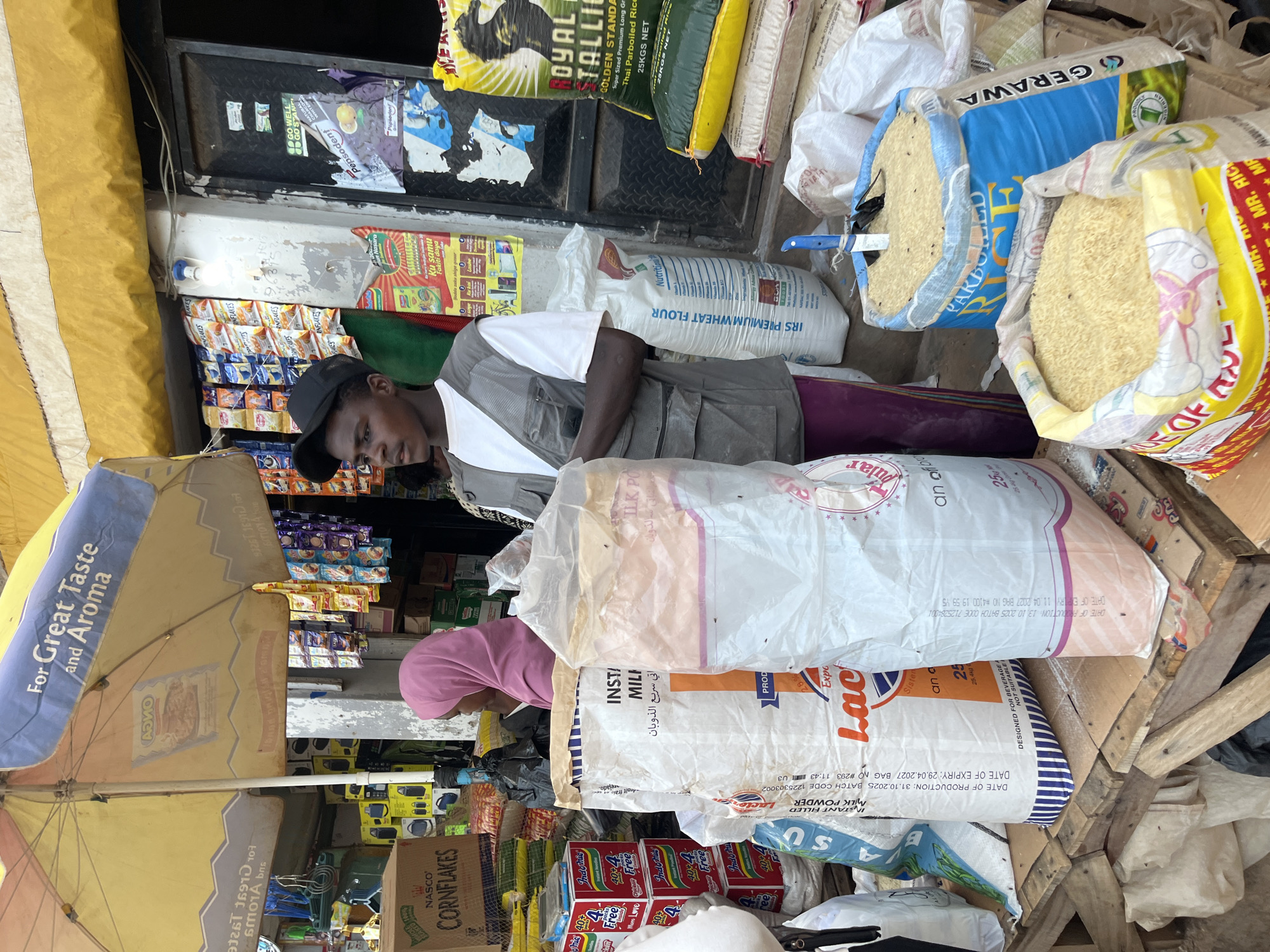 This is a medium shot, eye-level, outdoor photograph taken at a street market in Bukuru, Nigeria. In the foreground, a young Black male wearing a grey vest over a white t-shirt and a black baseball cap stands looking to the left of the frame. He is positioned behind a table covered with sacks of rice and flour. In the background, a woman wearing a pink hijab is partially visible to the left. Behind the people, the market stall displays an array of packaged food items, including boxes of cornflakes and instant noodles, hung from the ceiling and shelves. A large yellow umbrella with the words "For Great Taste and Aroma" is visible at the top of the frame. The overall environment appears to be a busy, bustling marketplace.