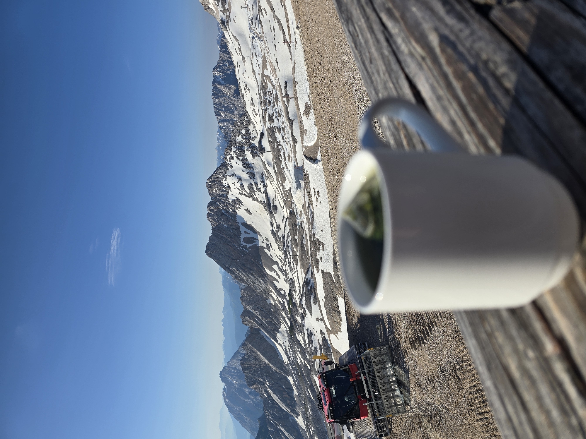 This is a photograph taken from a high vantage point overlooking a mountainous landscape. In the foreground, a white mug containing a green liquid, likely tea, is blurred out of focus, resting on a weathered wooden railing. Behind the mug, a red vehicle, appearing to be a snow groomer, is parked on a gravel surface. Beyond the vehicle, a vast expanse of snow-covered mountains stretches into the distance under a clear blue sky. The scene suggests a location in the Bavarian Alps, possibly near Garmisch-Partenkirchen, Germany, given the presence of the snow groomer and the alpine scenery, hinting at a ski resort or a high-altitude area. The lighting indicates it is daytime, with the sun casting shadows.