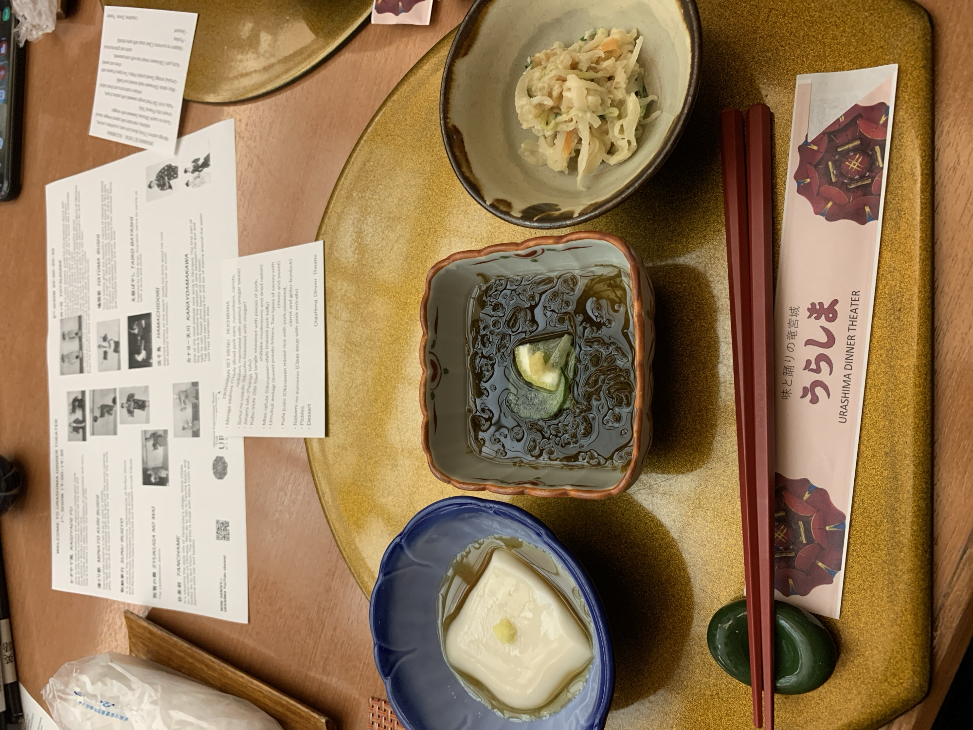 This is a close-up, eye-level, overhead shot of a table at the Urashima Dinner Theater in Naha, Japan. The scene is set at a table with a golden placemat. In the foreground, a pair of dark red chopsticks rests on a green chopstick rest. To the right of the chopsticks is a paper wrapper with a floral design and Japanese text that reads "Urashima Dinner Theater."
Three small dishes of food are arranged on the placemat. The dish on the left is a blue ceramic bowl containing a white, cube-shaped tofu with a small dollop of yellow paste on top, submerged in a clear broth. The center dish is a square, brown and white ceramic bowl filled with a dark, viscous liquid, a thin slice of green cucumber, and a piece of lemon or ginger. The dish on the right is a light brown ceramic bowl holding a pale salad, possibly coleslaw, mixed with shredded carrots and other vegetables.
In the background, a printed program or menu is visible, partially folded. It has Japanese text and several small black and white photographs depicting people in traditional attire. Some of the visible text on the menu includes "WELCOME TO URASHIMA DINNER THEATER" and "SHOW 19:00-20:30". A white plastic bag, possibly containing napkins or additional items, is on the left side of the frame, resting on a wooden surface. The overall impression is that of a well-presented meal at a cultural dining establishment.