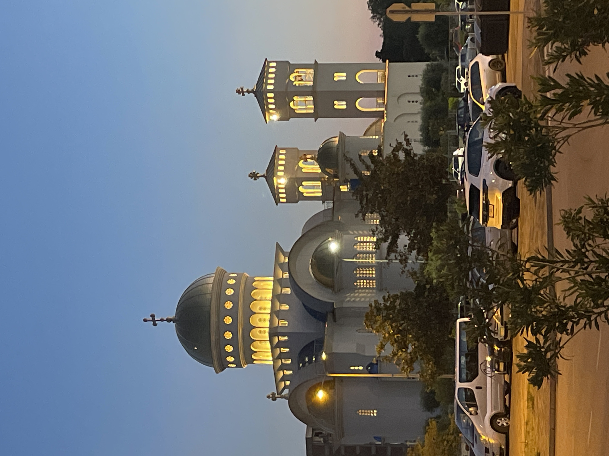The image displays a prominent Orthodox church in Bar, Montenegro, at dusk or early night, captured with a vertical orientation. The sky is a clear, deep blue, subtly transitioning to lighter shades on the right. The large, ornate church building is the central subject, featuring multiple domes and towers. The main dome, dark green and topped with a cross, is ringed with glowing, arched windows. Two smaller towers, also adorned with crosses, exhibit similar architectural details with illuminated windows, suggesting warm light within.
Along the right side of the frame, a street runs parallel to the church, lined with several parked cars, including white sedans and a white van, partially obscured by dark green trees and bushes. The street and pavement are illuminated by unseen light sources, casting a warm glow. No human activity is visible. The scene evokes a tranquil and majestic atmosphere, highlighting the church's architectural beauty against the twilight sky. No visible text is clearly discernible in the image.
